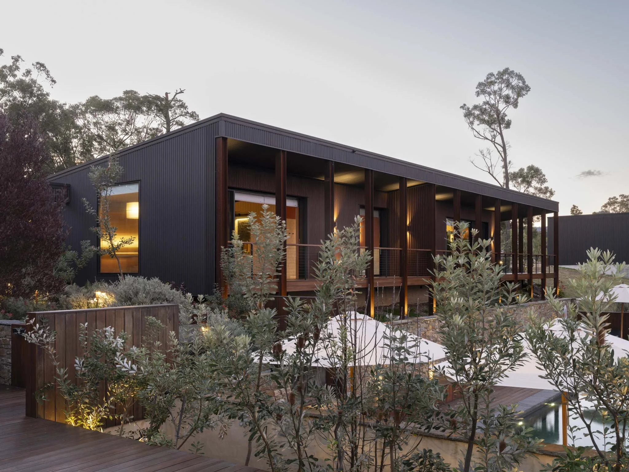 Modern black and corten steel architectural home with wooden accents, surrounded by lush plants, with lights on during dusk. #LuxuryTraveler #HiddenGem #SurroundedByNature