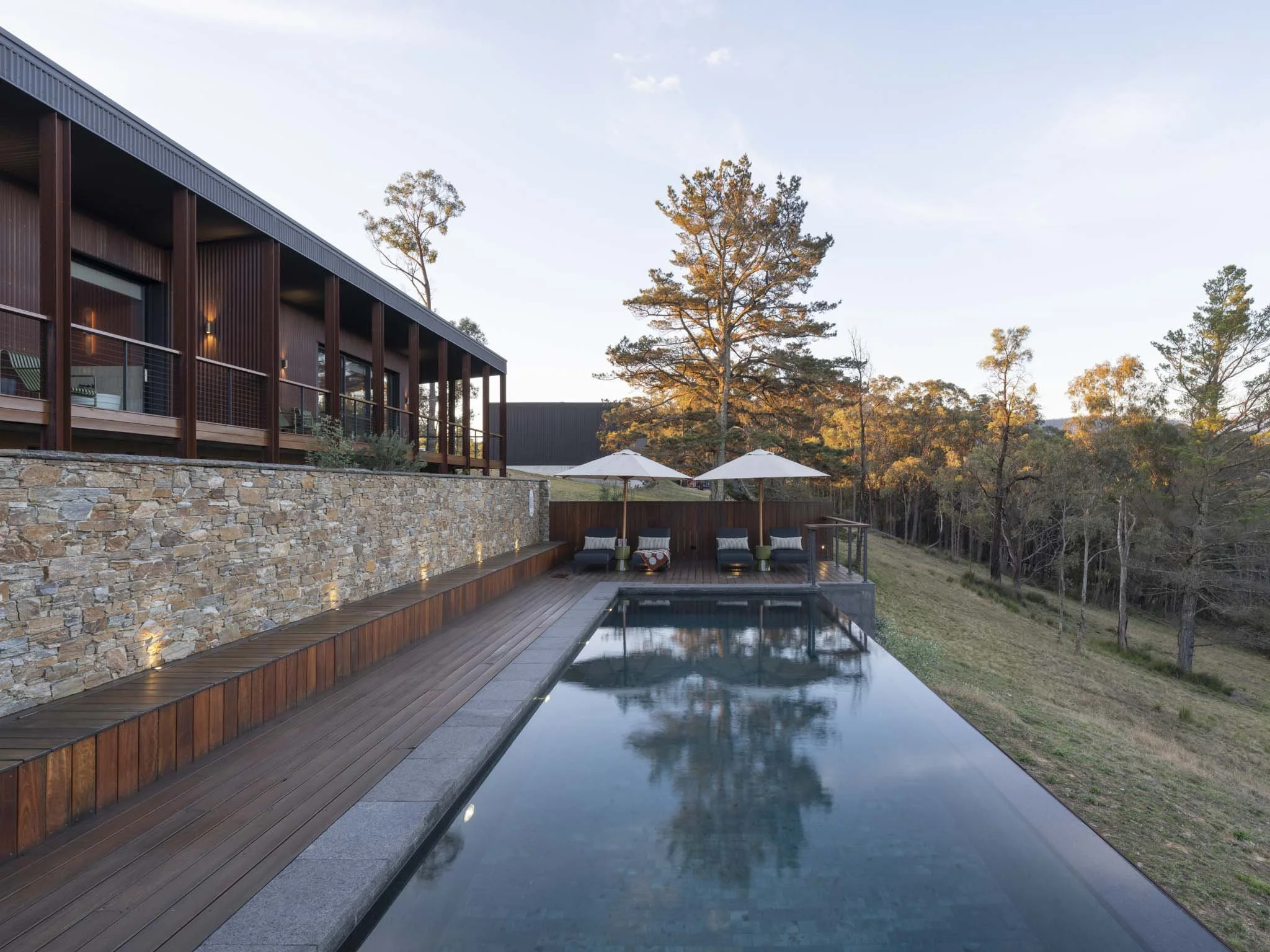 Modern house with outdoor pool and patio seating area, surrounded by trees and rolling hills at sunset.