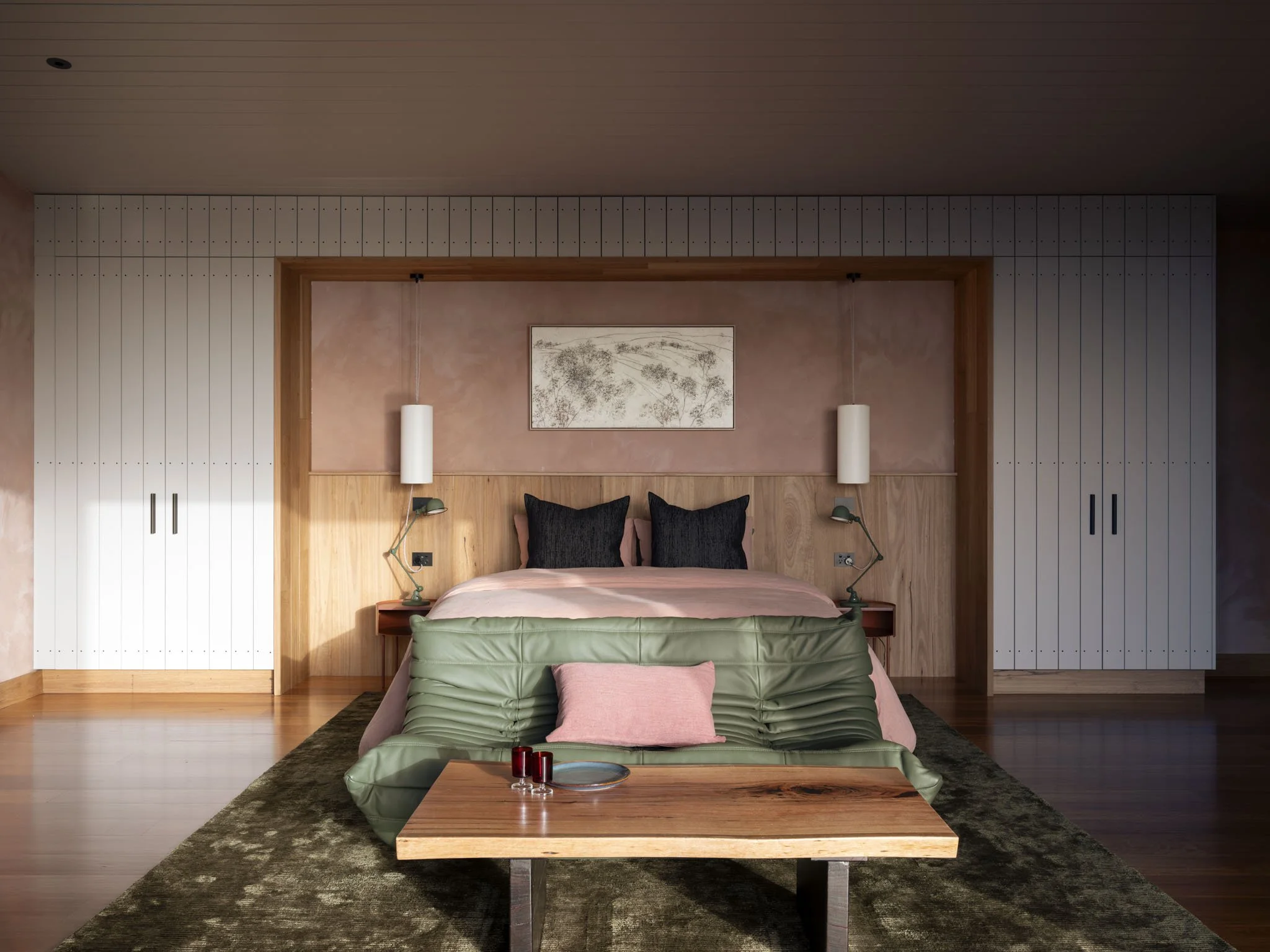 A modern designer bedroom with a king bed, native timber headboard, pink linen bedding, and black hand wovenpillows. There are two off white pendant lights above the bed, a hand made blackbutt slab coffee table with two vintage red glasses, and a fra