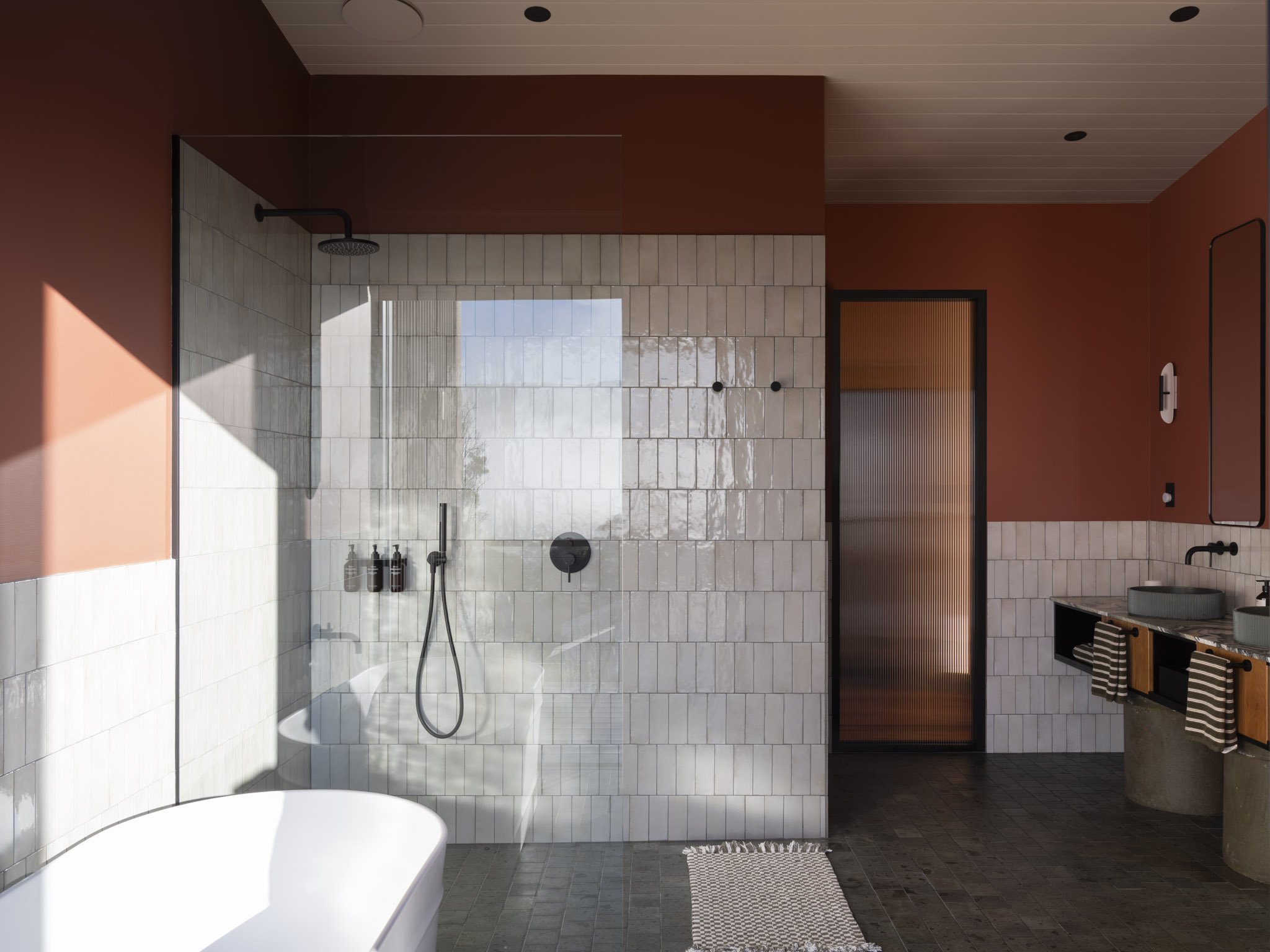 Modern rustic luxury spa bathroom with a large walk-in shower featuring white subway tiles, a rain showerhead, freestanding soaking bathtub and black fixtures. The bathroom has earthy tones with dark wood and black accents, a double vanity with flute
