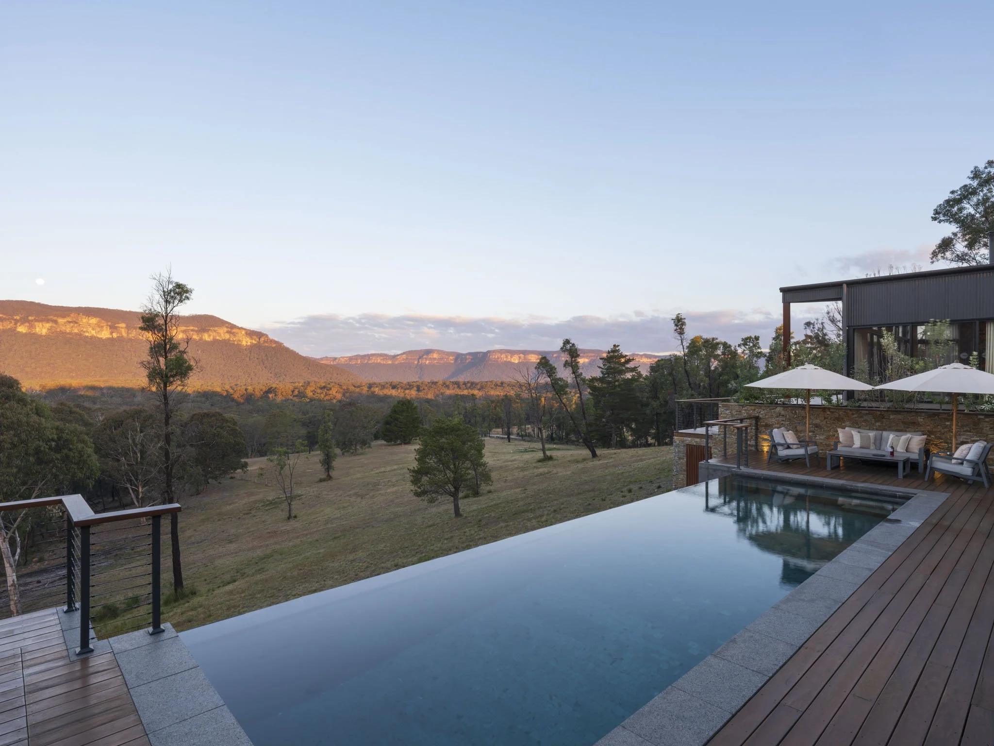 Outdoor infinity heated swimming pool on a wooden deck with relaxing seating area, overlooking a landscape of native bushland and Blue Mountains ridgeline during sunset. #RomanticGetaway #SydneyStaycation #Travelgoals #WanderLust #FiveSongs.au