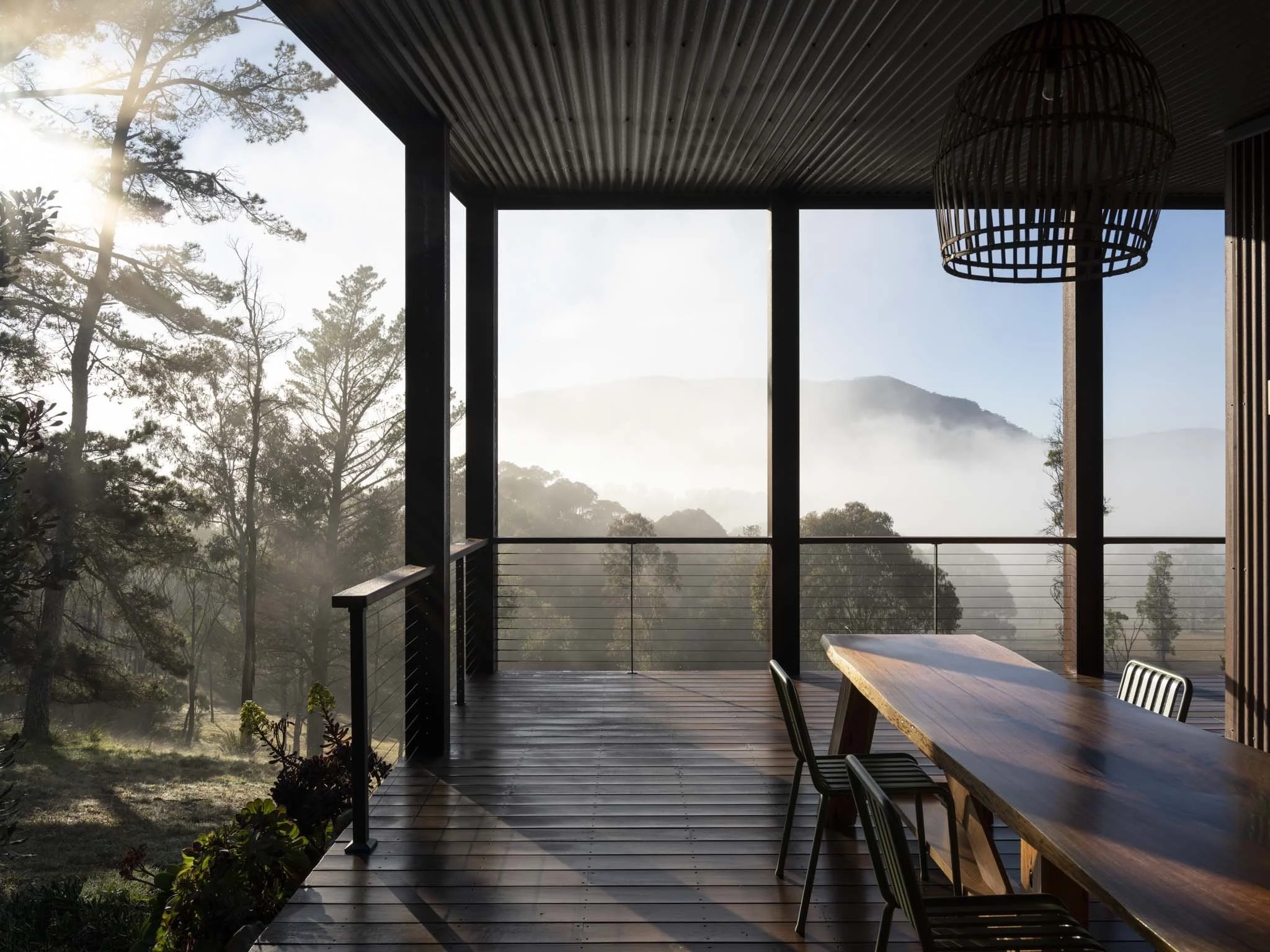 A spotted gum deck with a hand hewn blackbutt timber slab dining table and designer chairs overlooks a misty Megalong Valley landscape, with native bushland and Blue Mountains in the distance and the sun shining through the branches. #SpectacularView