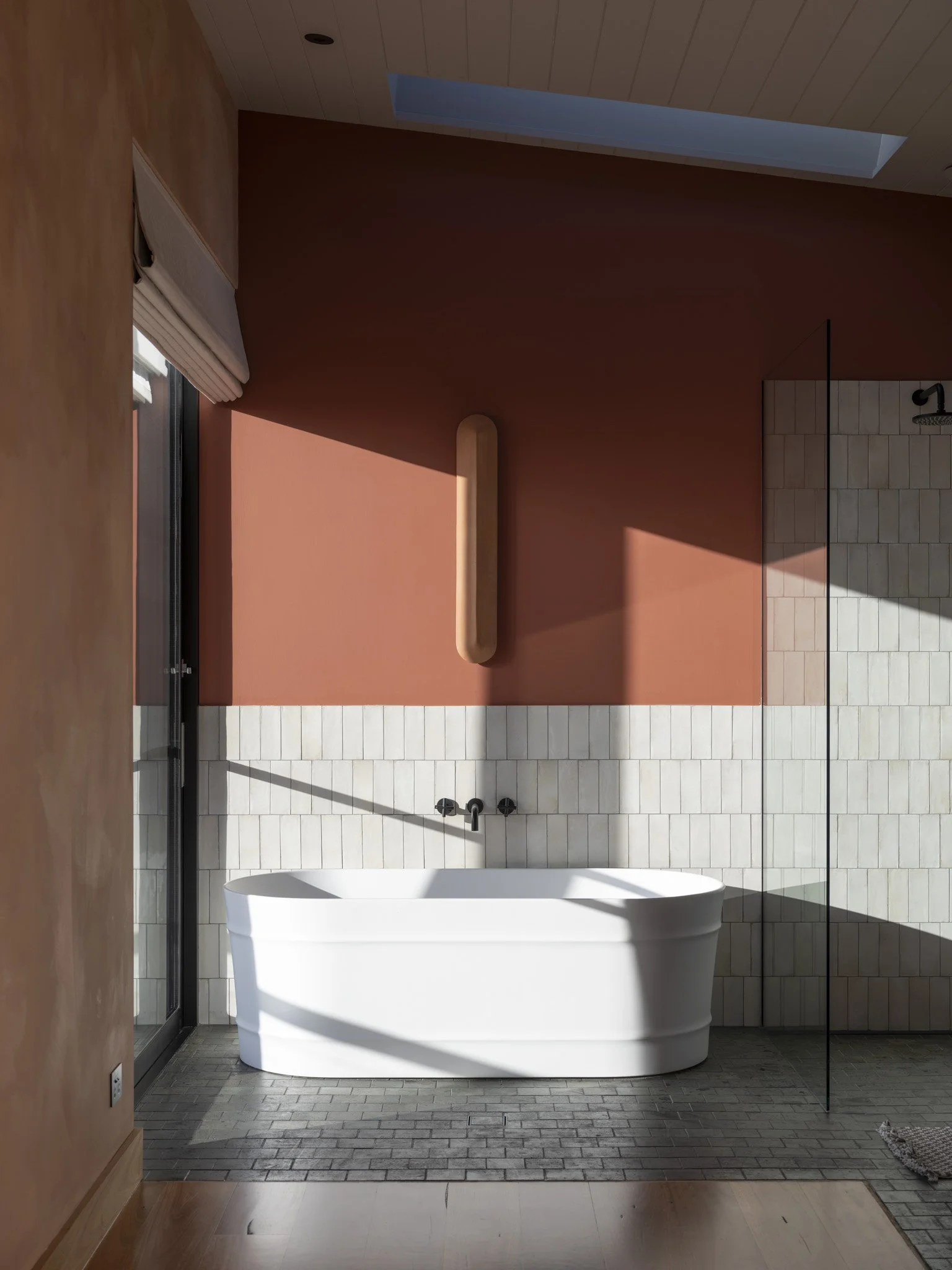 Modern designer bathroom with a white freestanding bathtub, a wall-mounted black faucet, a terracotta-colored accent wall, a wooden panel in the shower area, a skylight, and floor-to-ceiling glass shower enclosure. #StaycationGoals #WeekendAway