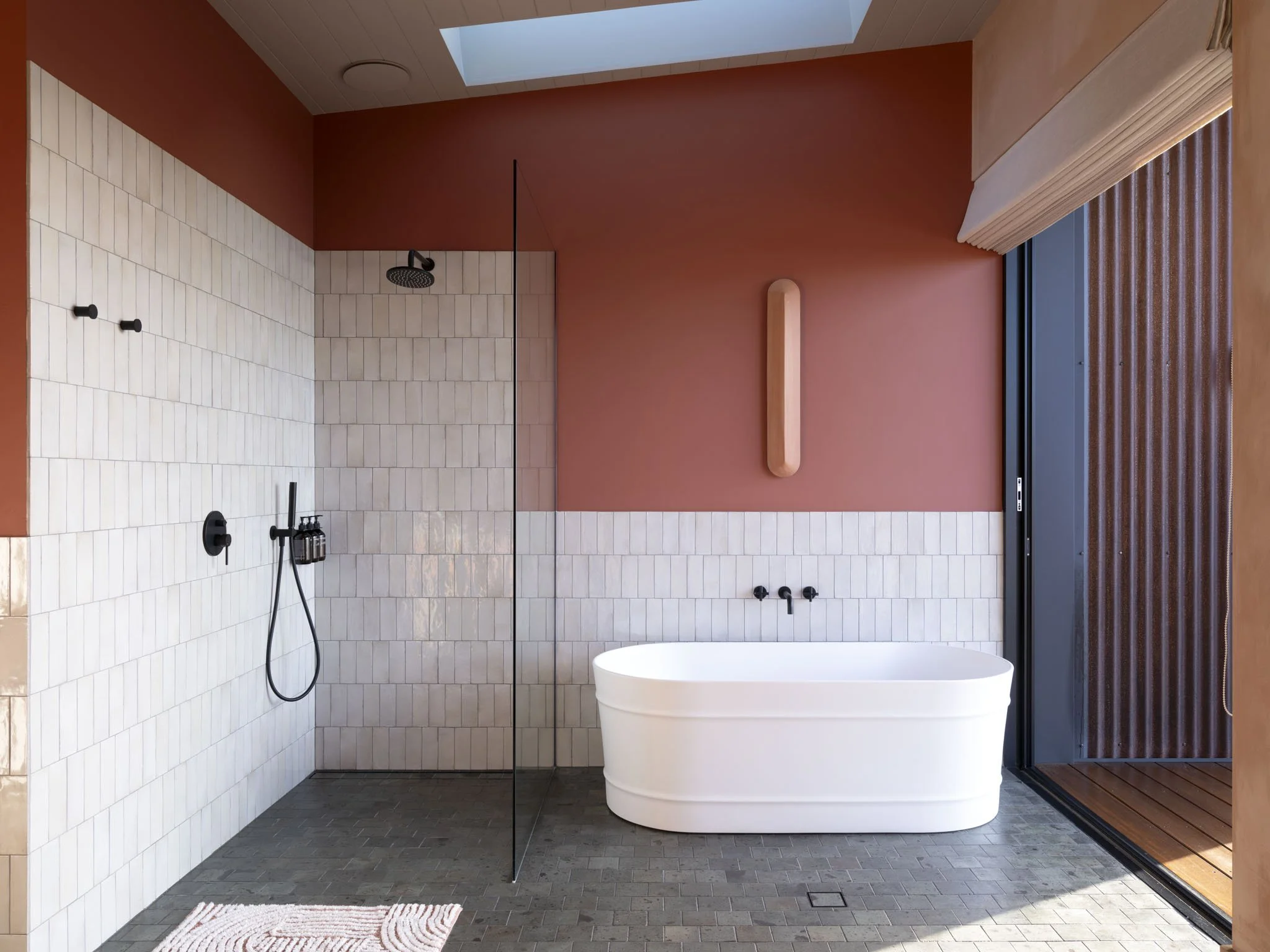 Modern luxury spa bathroom featuring a walk-in shower area with a black rainfall showerhead and handheld shower, freestanding soaking bath, terracotta designer lamp, large window with privacy blinds, and a geometric patterned rug on the natural stone