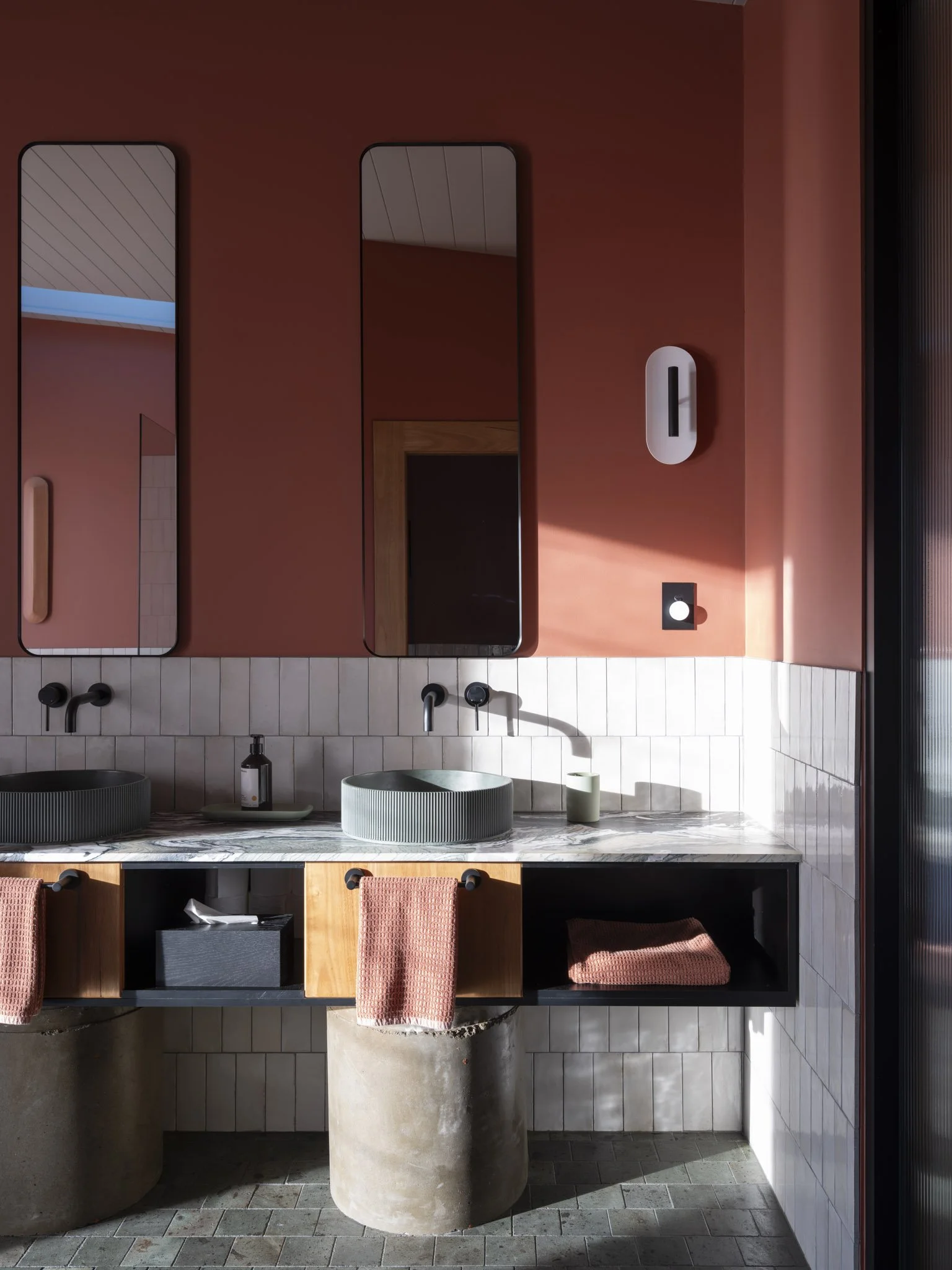 Modern designer bathroom with a double fluted concrete sink, black fixtures, and a large marble countertop. Pink tiled wall with black-framed mirrors. Wooden open storage beneath the countertop with pink towels and a black box. #RusticLuxury