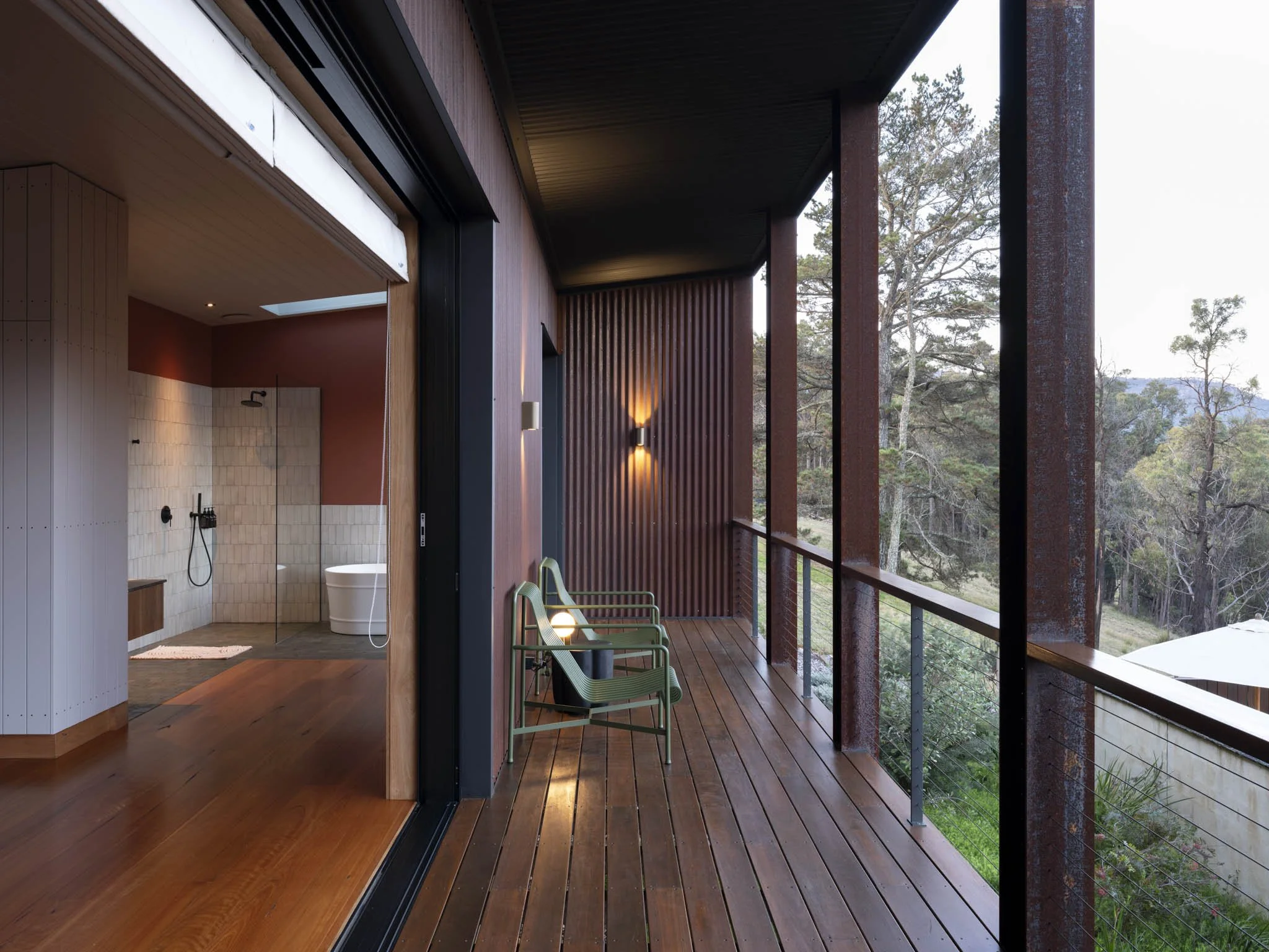 The image shows a modern outdoor balcony with wooden flooring and a railing, overlooking a forested landscape with trees and a distant sky. There are two green designer lounge chairs on the balcony, and the setting appears to be during daytime. #Stay