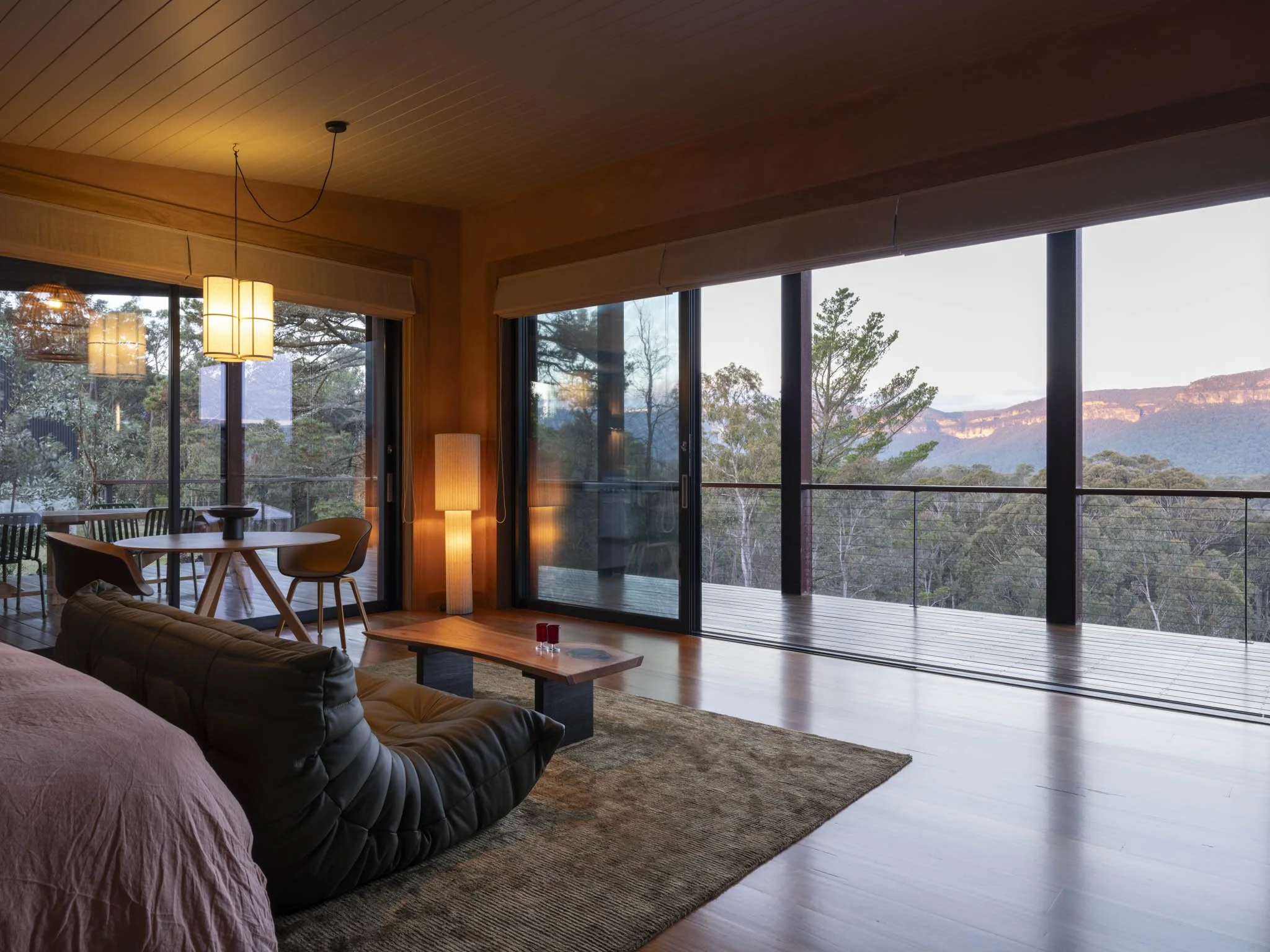Rustic luxury living and bedroom with large sliding windows showing a spectacular Blue Mountains view, some designer chairs, a table, and a tall floor lamp. An olive green leather sofa and green bamboo silk rug to ground the space. #SpectacularViews 