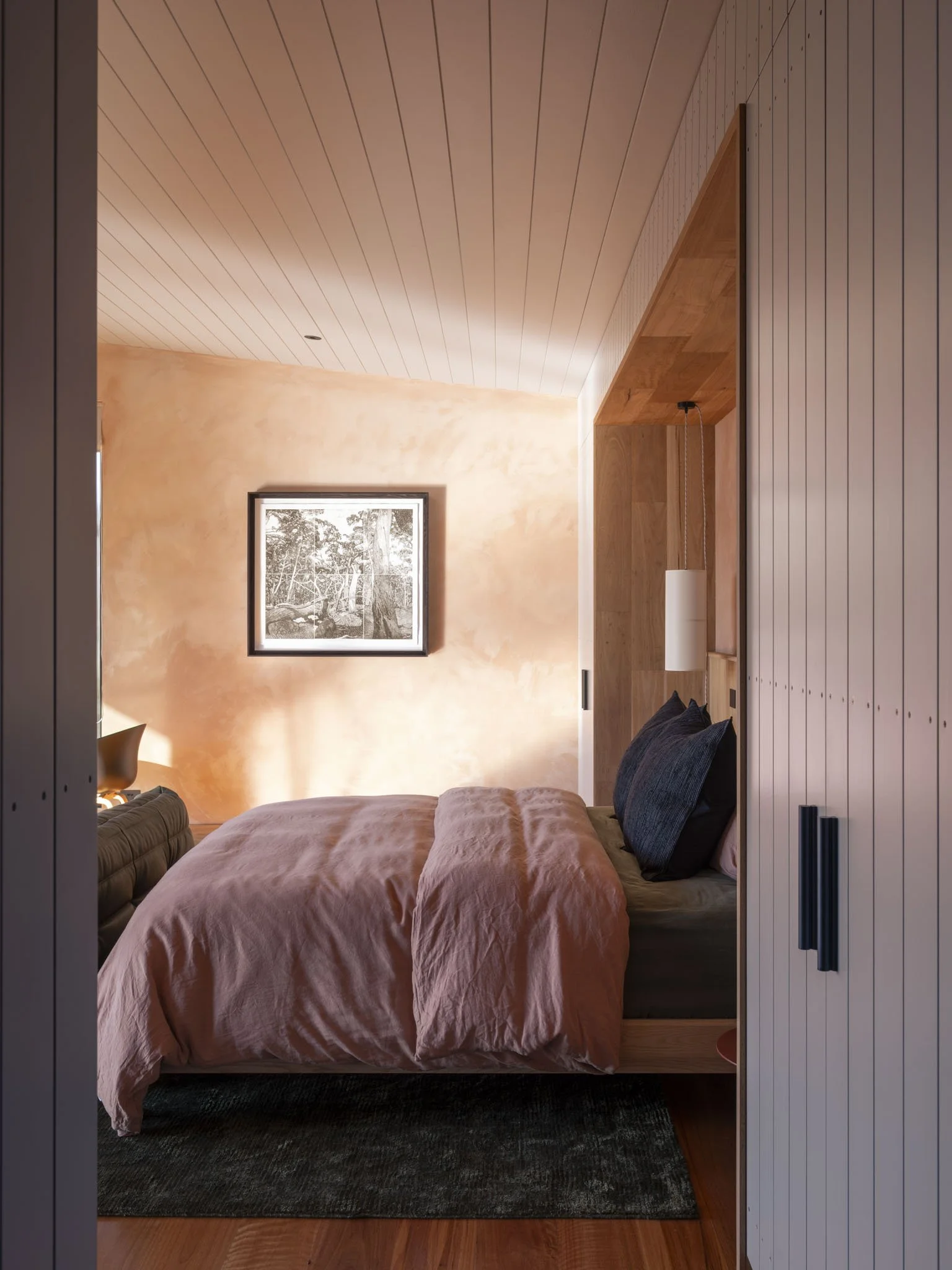 A bedroom with a pink linen bed cover, hand woven pillows, and a black and white framed artist's etching on a lime-washed terracotta wall. There is a built-in native timber headboard and a hanging pendant light. #RomanticGetaway #RusticLuxury