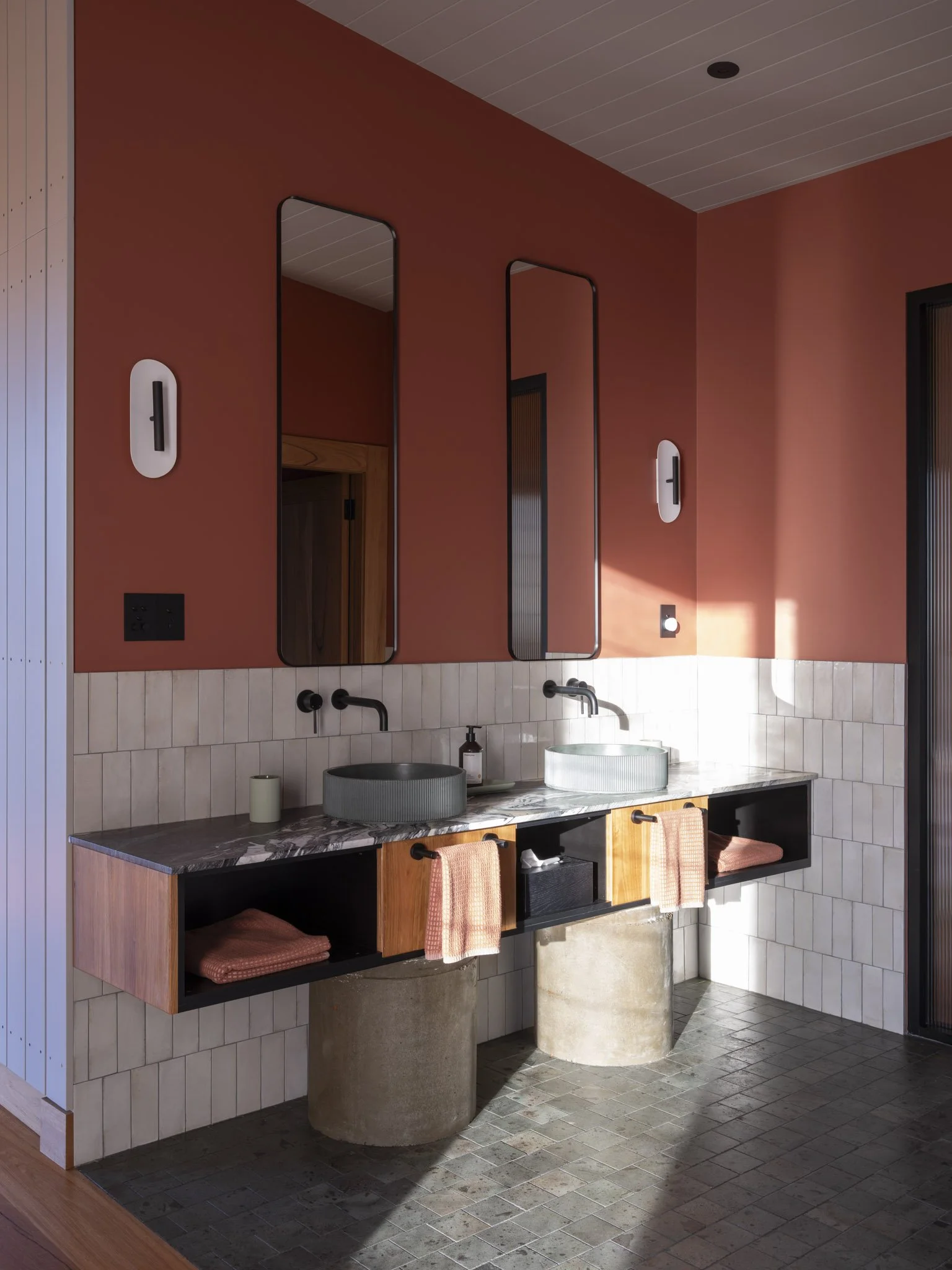 Bathroom with double vanity and fluted green concrete sinks on a black marble countertop, two tall mirrors, modern australian designer wall-mounted light fixtures, and a mix of natural wood, stone and concrete accents.