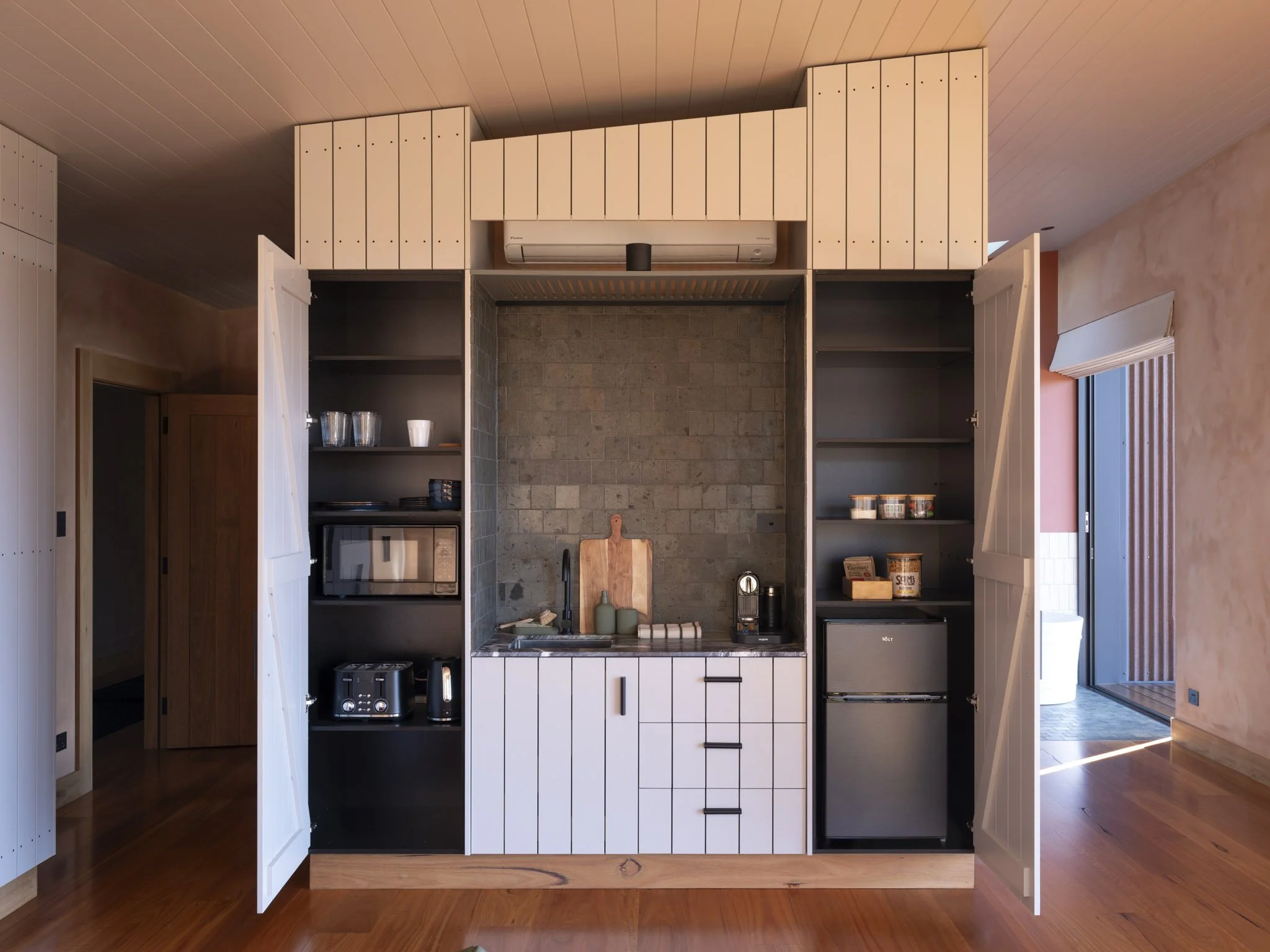 Kitchenette cabinet with open doors, showcasing shelves with various kitchen items, and a microwave oven, refrigerator at the bottom. Natural stone tiles and marble countertop. #StaycationGoals #WeekendAway #SlowLiving #SlowMornings