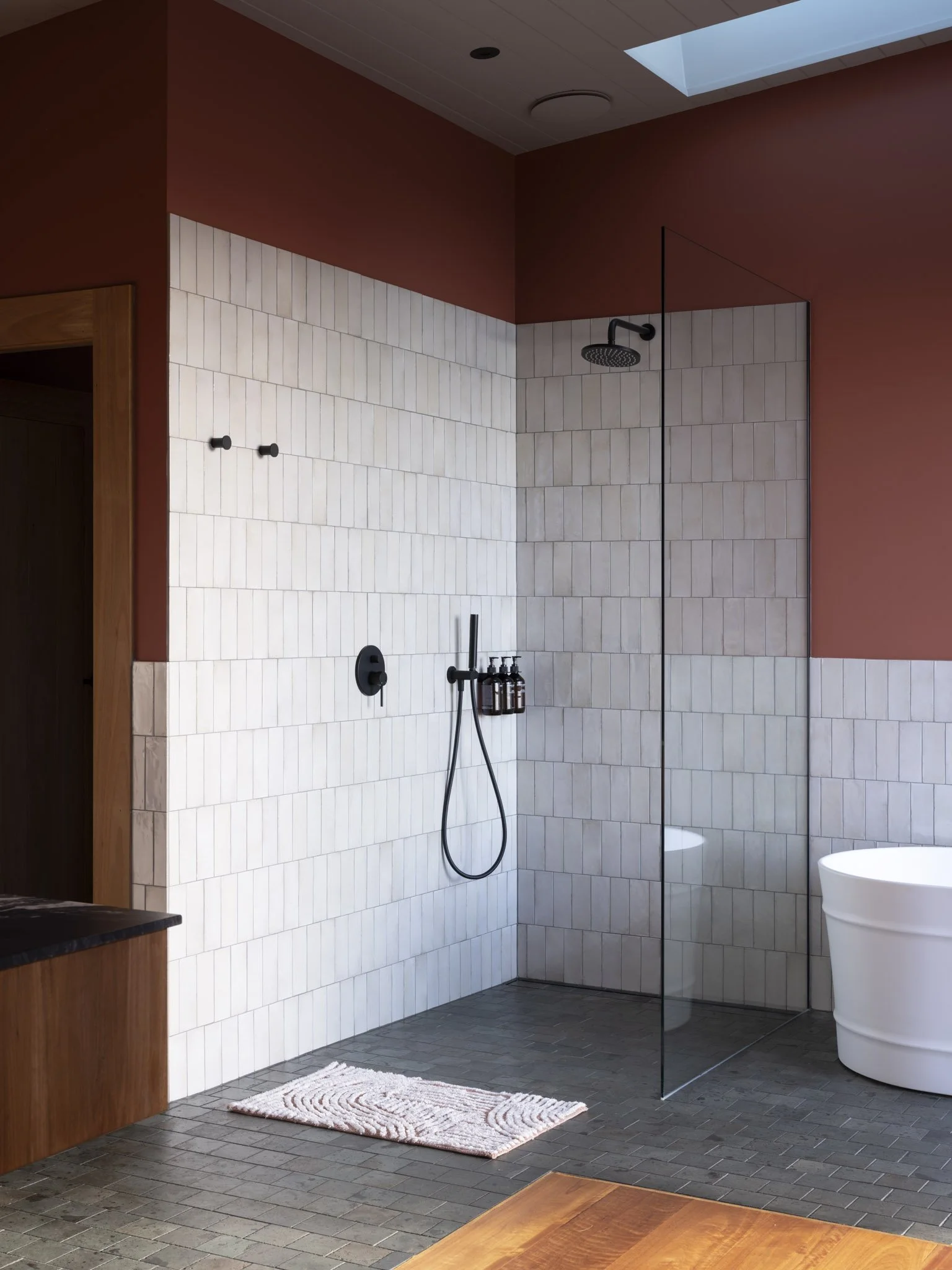 Modern bathroom with a walk-in shower area, white tiled wall, black fixtures, and a clear glass partition; a white freestanding bathtub, a patterned rug on gray tiled floor, and a window allowing natural light.