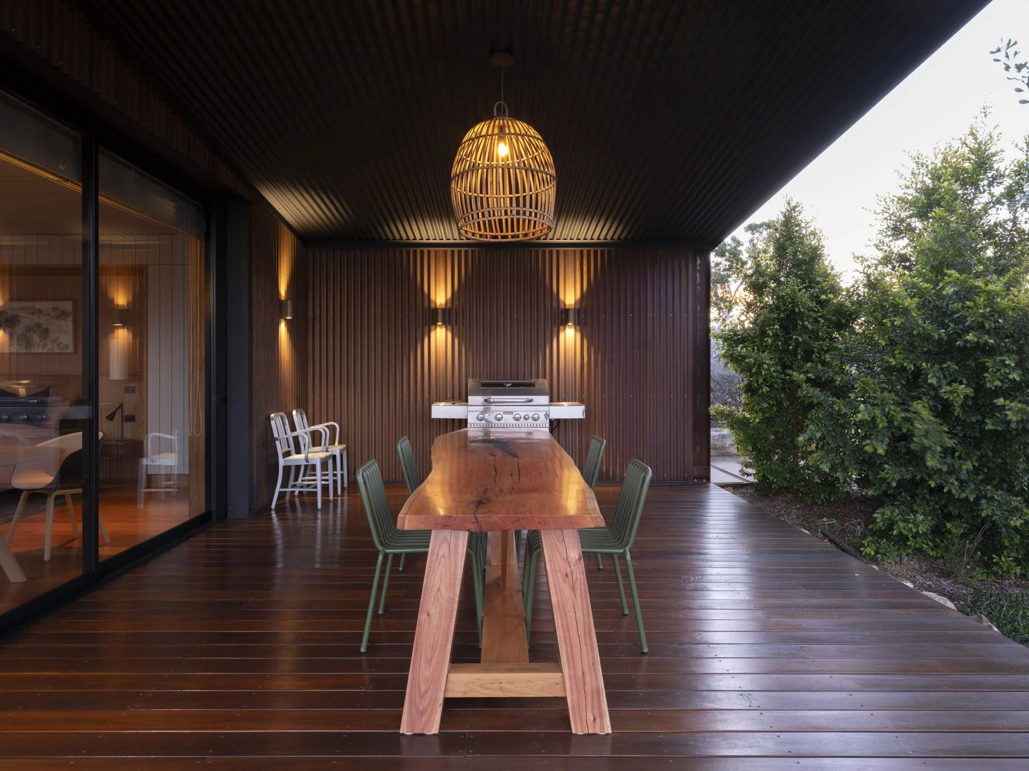Outdoor patio at the Evening Song guest room of the Five Songs rural retreat, with a long handmade blackbutt timber slab dining table, green designer chairs, a large barbecue grill, and modern lighting on a corten steel corrugated wall, with native t