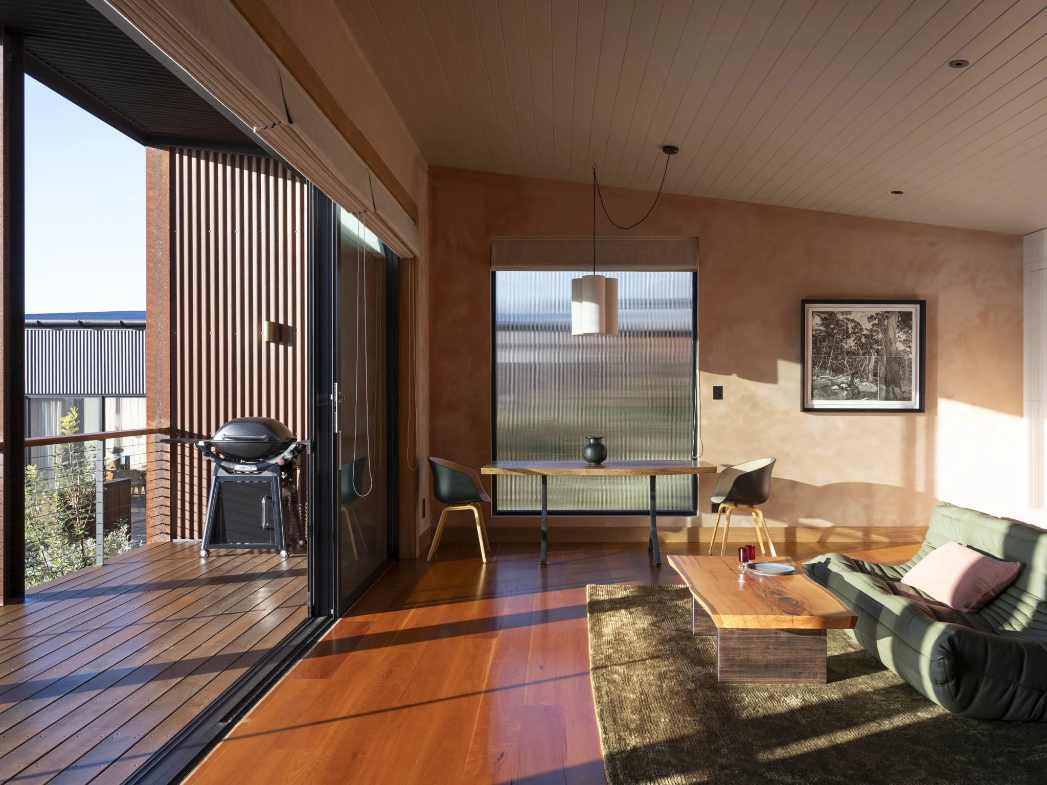 Modern rustic luxury living room with native timber flooring and ceiling, large windows, a hand made timber slab dining table with chairs, a green leather sofa, and an outdoor patio with a barbecue grill. #RomanticGetaway #RoomWithAView