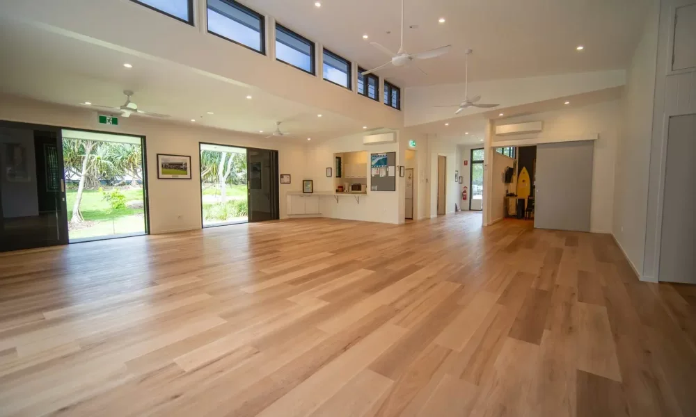 Spacious indoor area with wooden flooring, white walls, ceiling fans, and large glass sliding doors leading to an outdoor green space with trees.