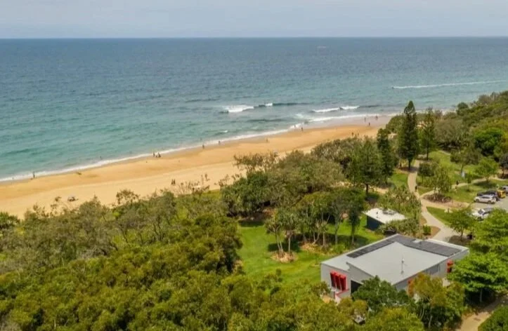 Aerial view of a beach with sandy shoreline, ocean waves, and lush green trees and park area with a building in the foreground.