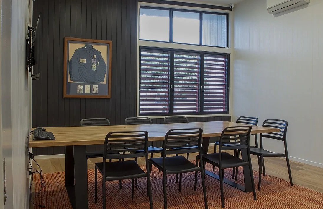 Empty conference room with a long wooden table, six black chairs, a framed photo of a police jacket on a dark wall, a window with shades, and a wall-mounted television.