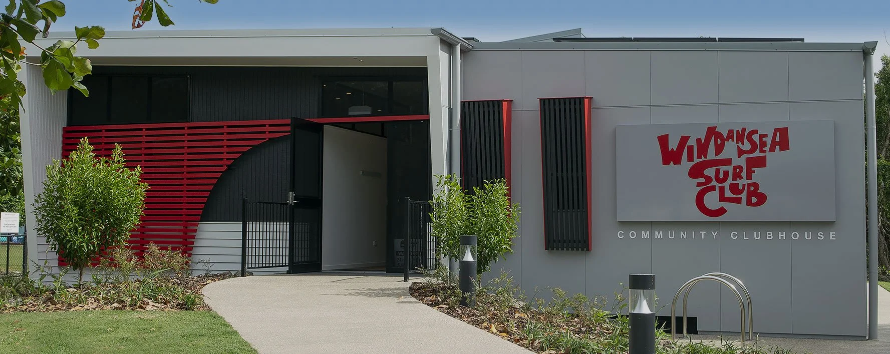 The WindanSea Surf Club Community Clubhouse with a modern exterior, featuring black and red architectural elements, surrounded by greenery, with a pathway leading to the entrance.