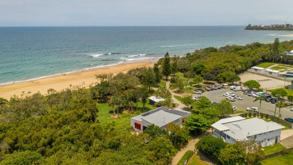 Aerial view of a beach with a sandy shoreline, ocean waves, and a parking lot surrounded by trees and residential buildings.