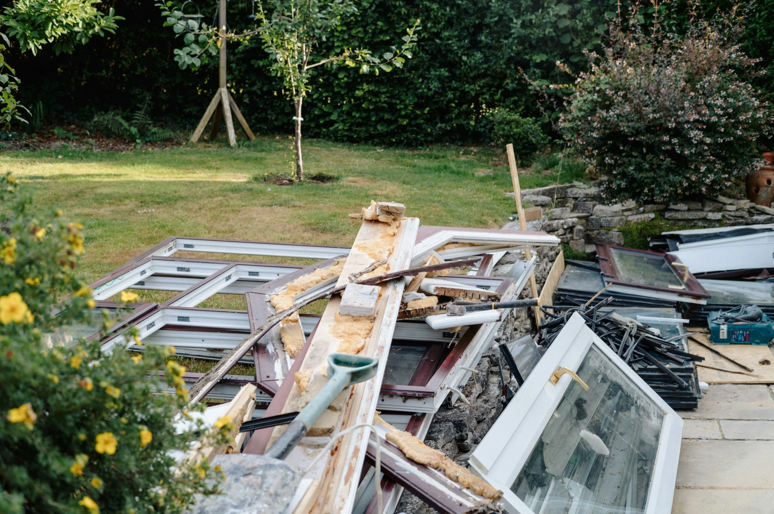A backyard with a pile of window frames, construction debris, and insulation material, surrounded by greenery, trees, and bushes.