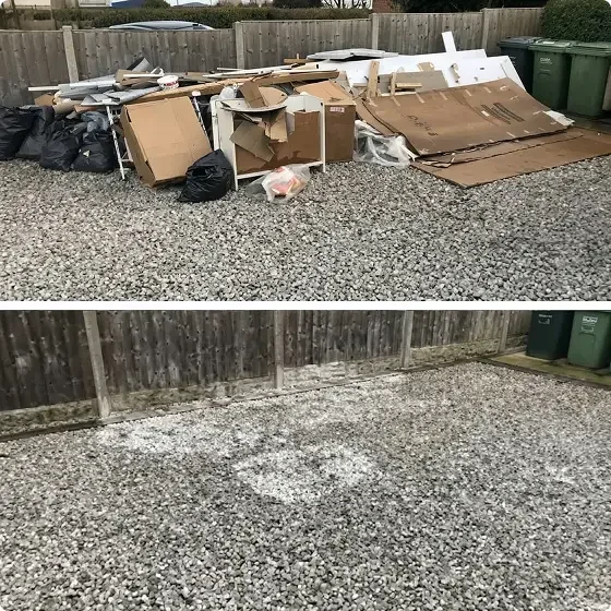Piles of discarded cardboard, paper, and trash bags on a gravel surface, with a wooden fence and green trash bins in the background.