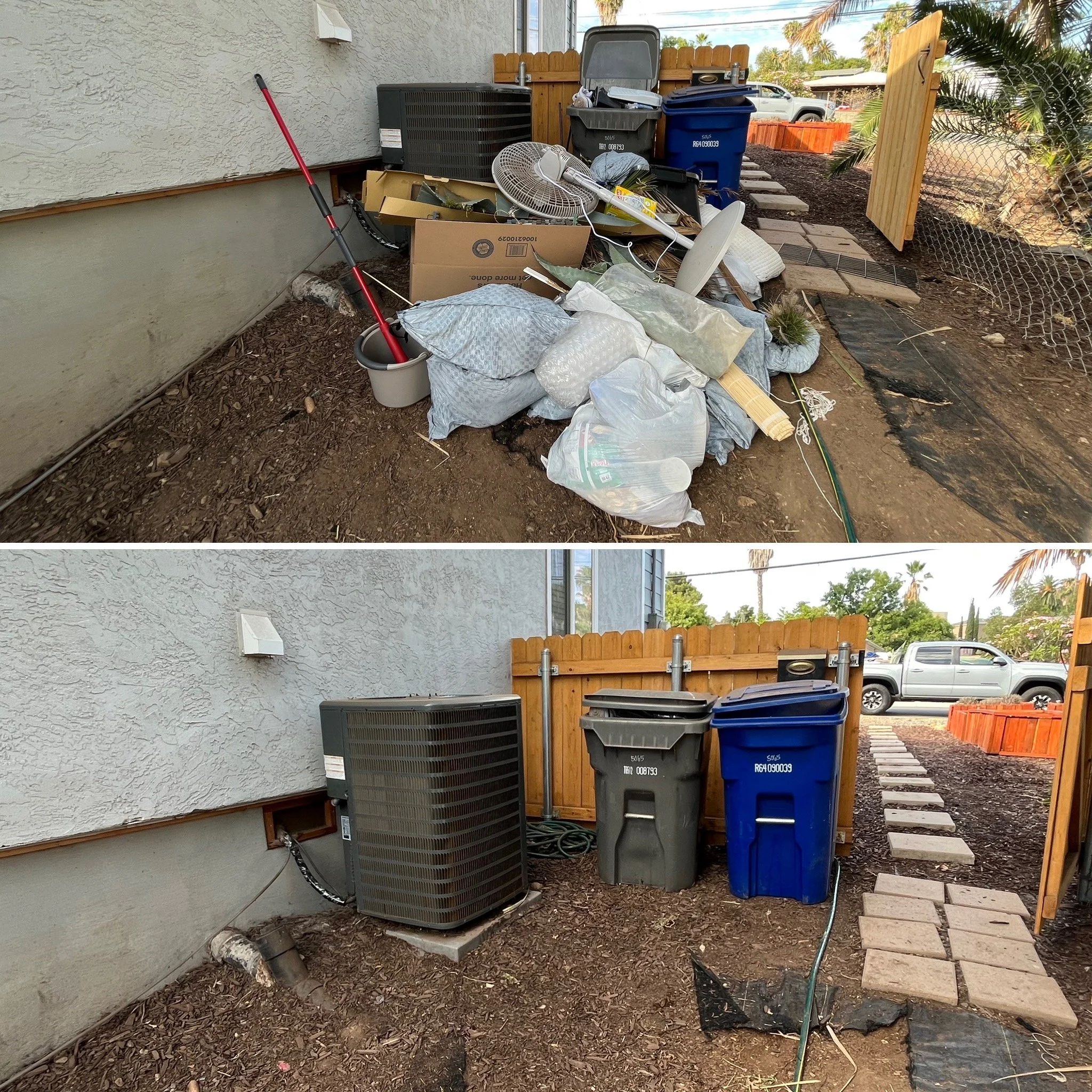 A cluttered backyard corner with garbage and recycling bins, discarded items, and a wooden fence with a gate. The ground is covered with dirt and stepping stones, and part of a house with a textured wall is visible.