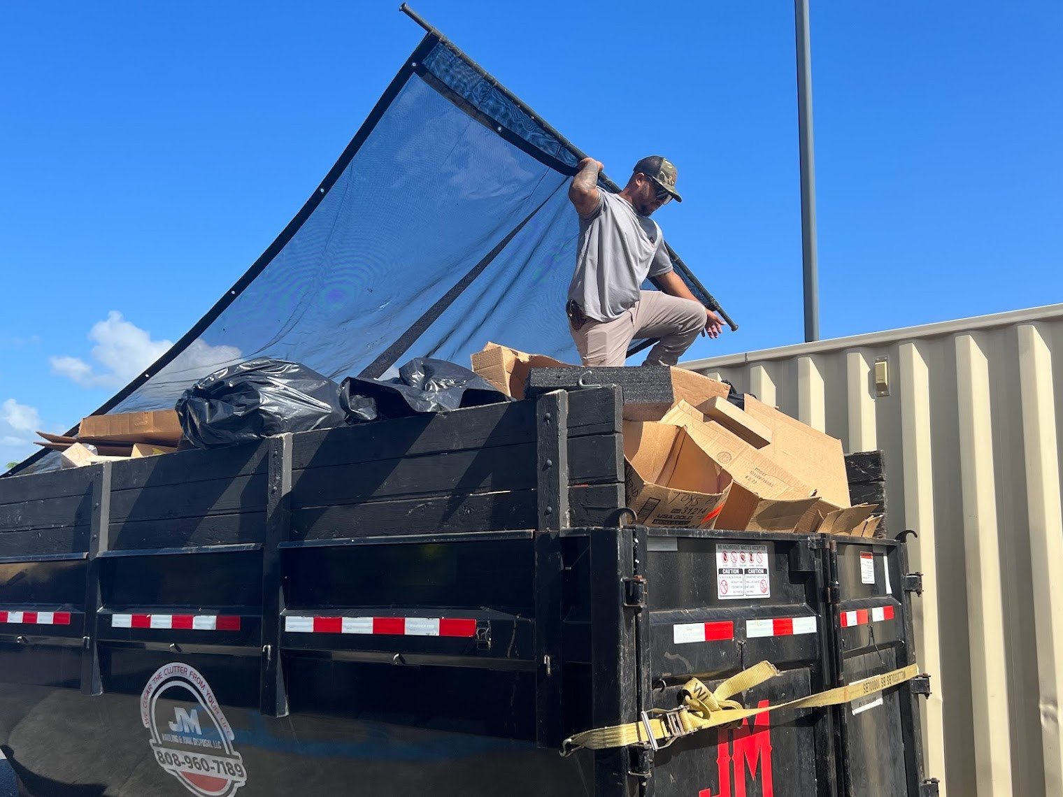 A worker standing on top of a black trailer filled with cardboard boxes and black trash bags. The worker is holding a large metal rod or pole, wearing a gray shirt, tan pants, and a camouflage cap. The background features a blue sky with clouds and a beige building.
