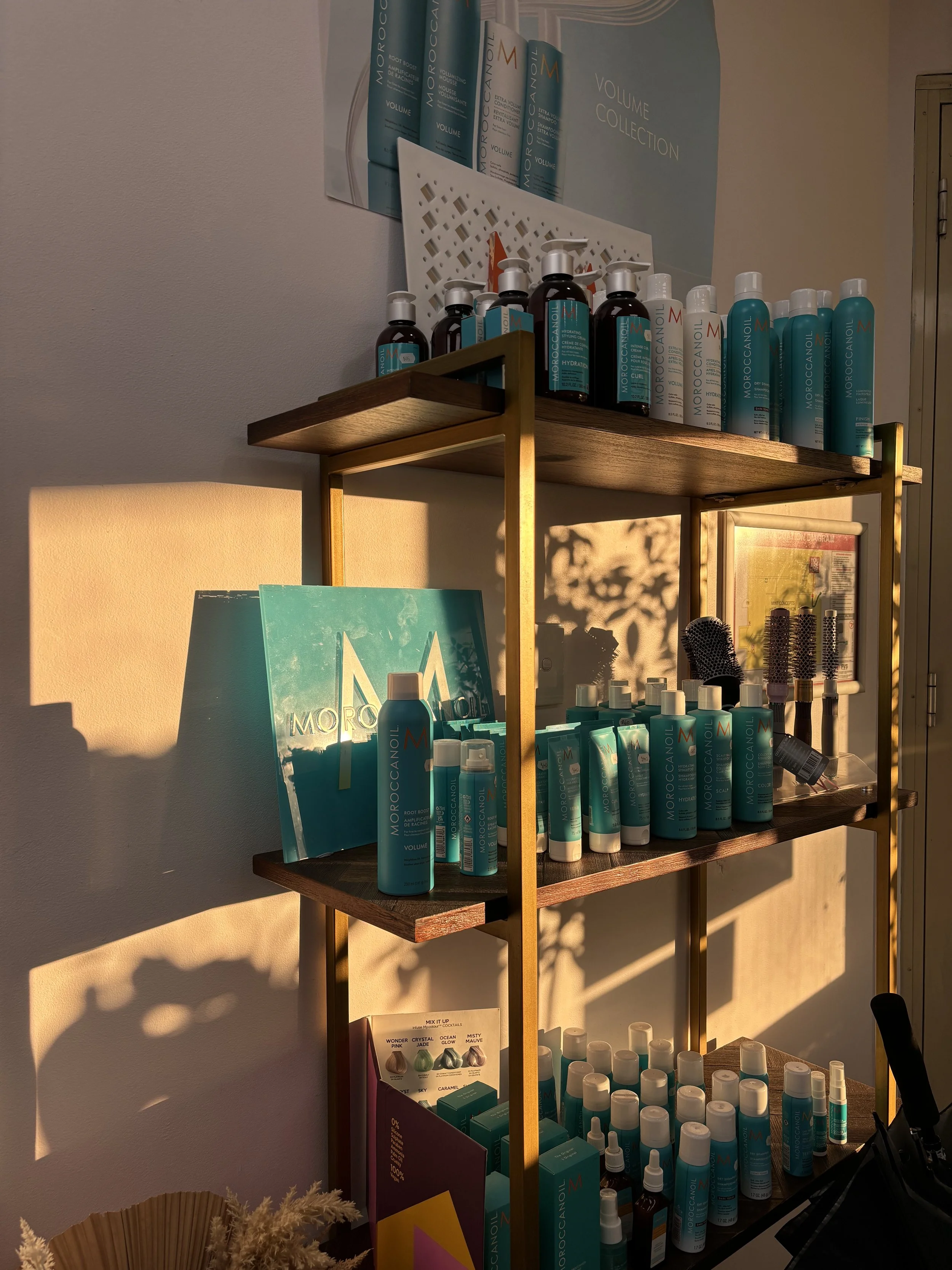 Display of Moroccanoil hair care products on a wooden and metal shelf, including shampoos, conditioners, and styling treatments, with a poster on the wall above and shadows cast on the wall and shelves.