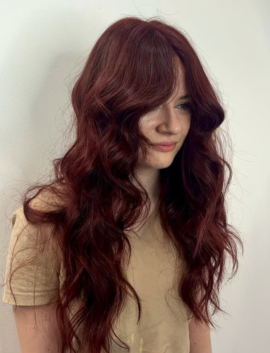 Woman with long, wavy, burgundy hair, wearing a beige top, looking down slightly, standing against a plain white wall.