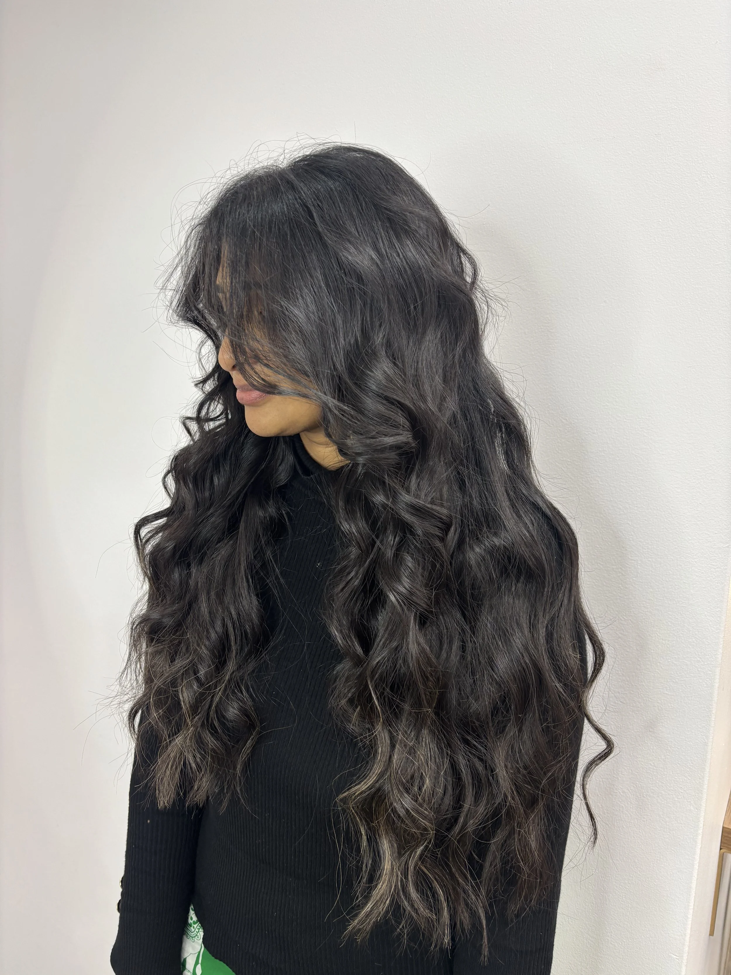 Woman with long, wavy dark hair with lighter highlights, wearing a black top, standing against a plain white wall.