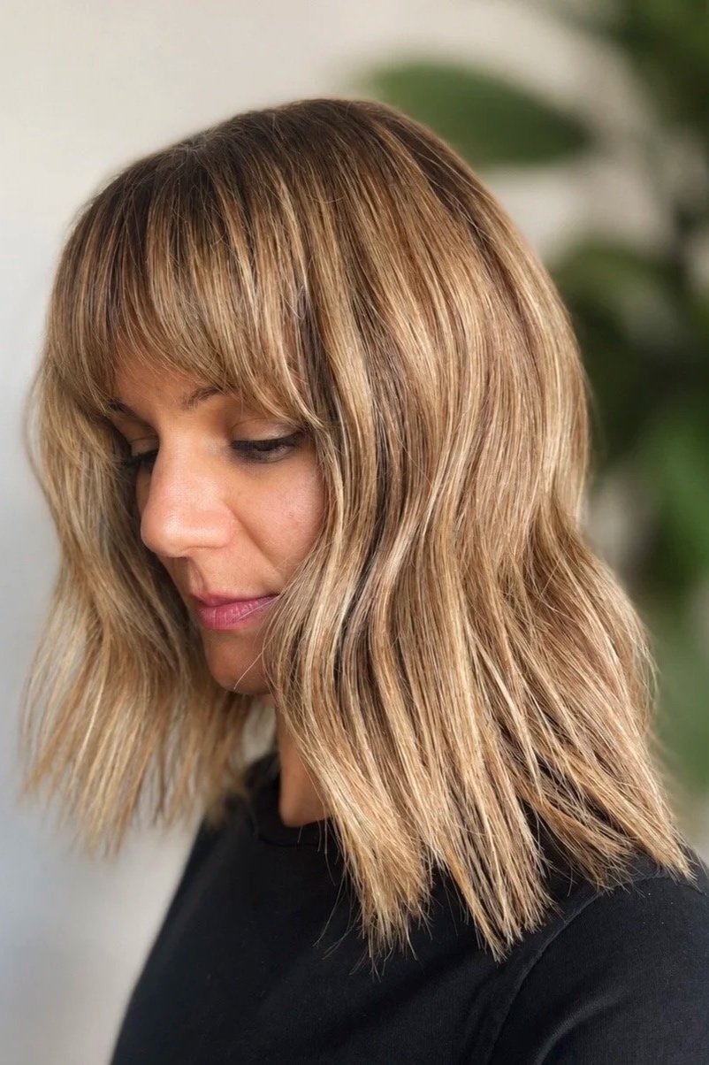 Close-up of a woman with shoulder-length wavy blond hair and bangs, looking downward, wearing a black top.