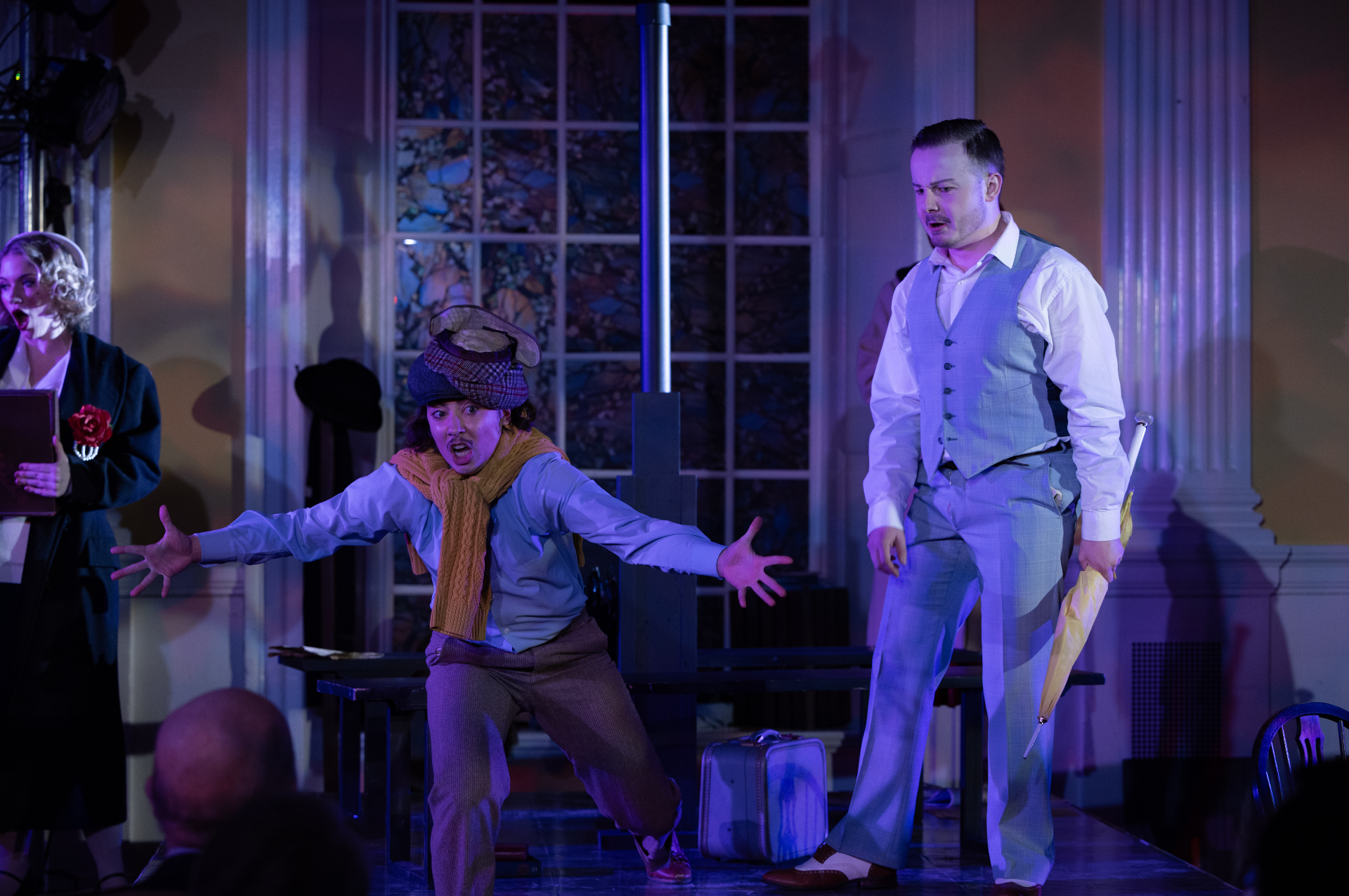 Actors performing a theatrical scene indoors, with one actor making a dramatic gesture, another dressed in a light gray suit with an umbrella, and a woman in the background holding a book, all under stage lighting.