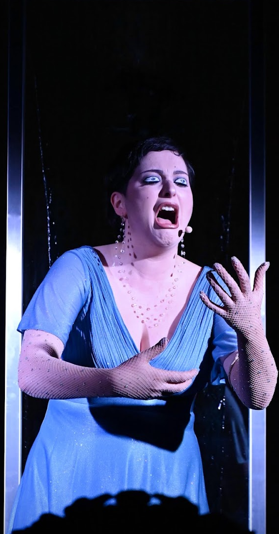 A woman with short dark hair wearing a blue dress and long glittery gloves, appearing to sing or shout intensely on stage, with a dark background and dramatic lighting.
