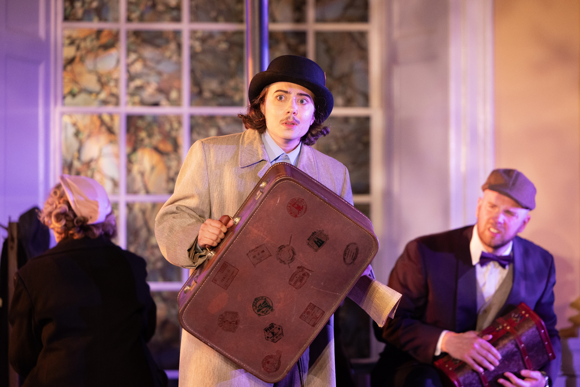 A person in a vintage brown coat and black hat holding a large suitcase, with a surprised expression, sitting inside a room with large windows and other people dressed in vintage clothing.