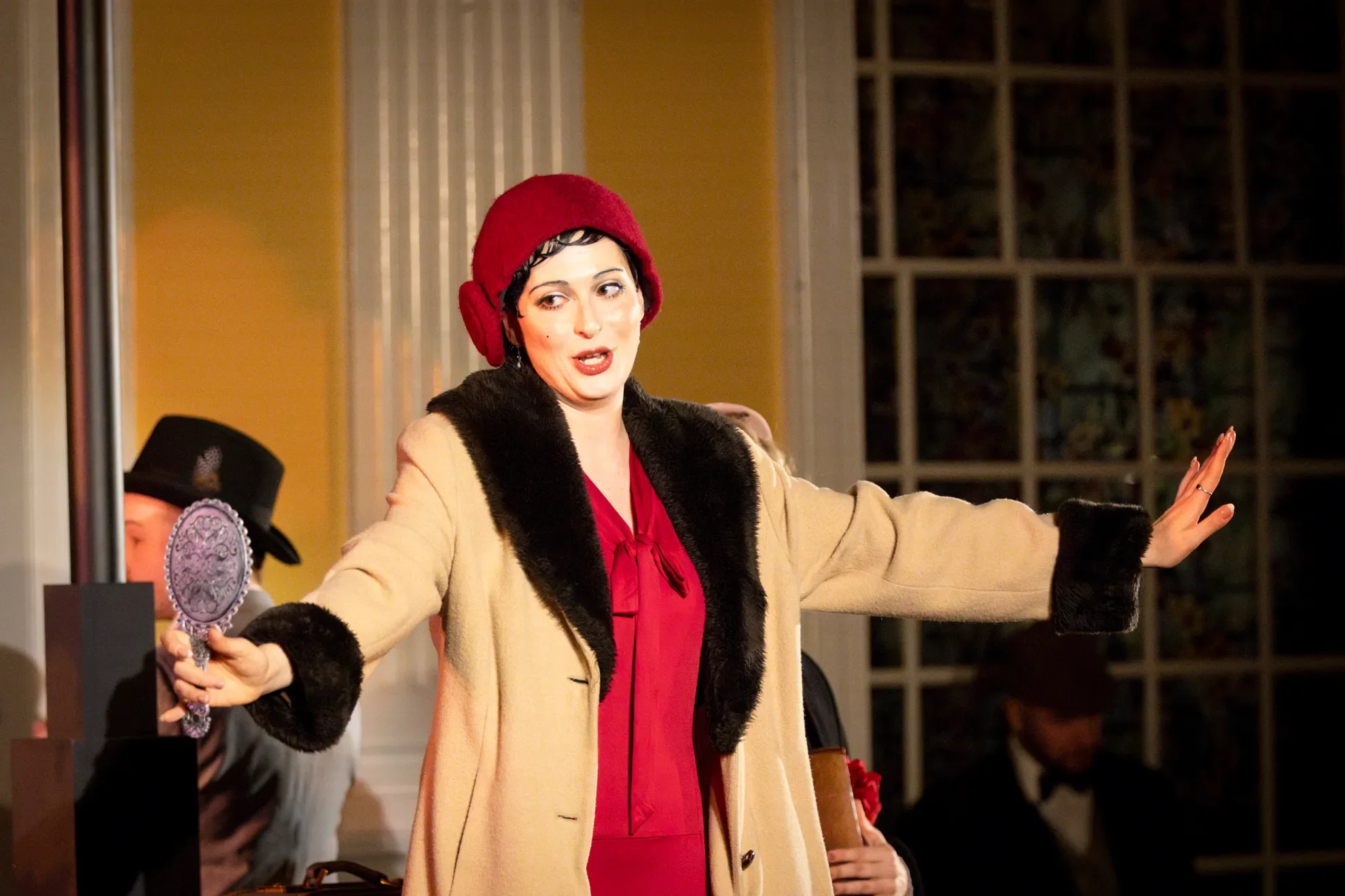 A woman dressed in vintage clothing with a red hat and beige coat with black fur trim, holding a hand mirror and gesturing with her other arm, appears to be on a stage or set with a yellow wall and windows in the background.