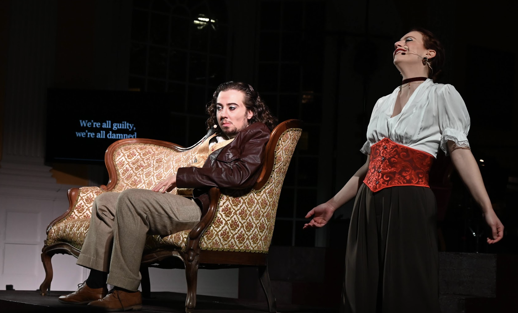 Two actors performing on stage, one seated on an ornate chair with a guitar, looking serious; the other standing with arms open, smiling. A screen in the background displays the words "We're all guilty, we're all damned."