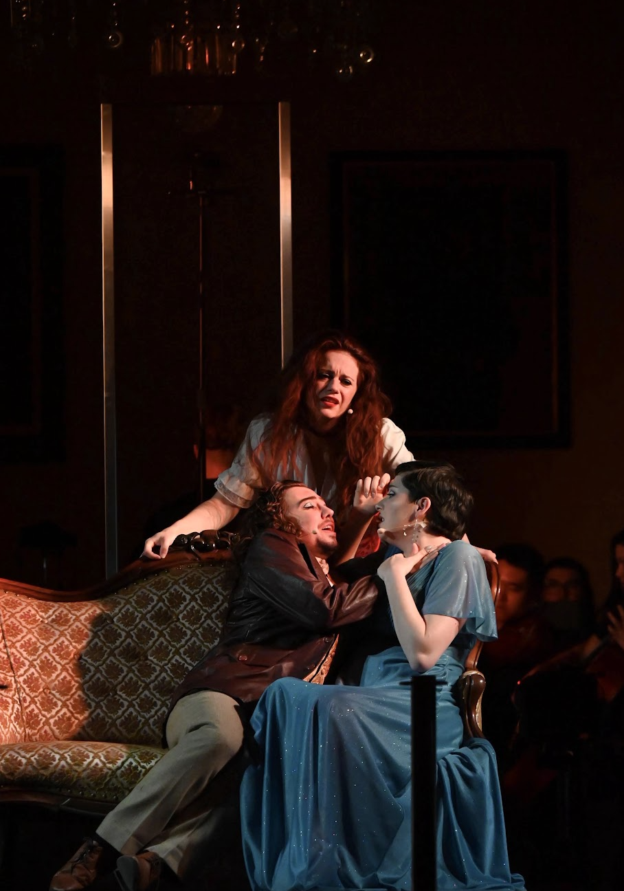 Three actors performing a dramatic scene on stage, with one woman standing behind two seated women, all dressed in period costumes.