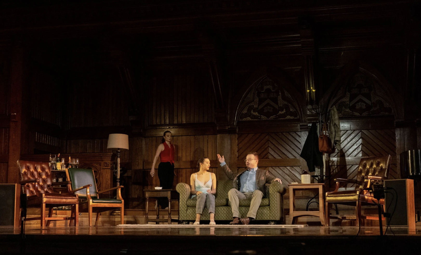A stage scene with three actors, one seated on a green couch, one standing behind it, and one sitting on a wooden chair nearby, in a room with wood-paneled walls and various furniture and objects.