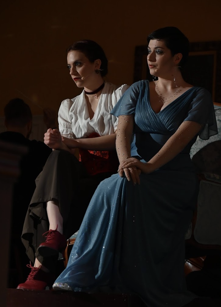 Two women sit together on a stage, one with short dark hair in a blue dress and the other with short brown hair in a white shirt and dark pants, watching an event in a dimly lit room.