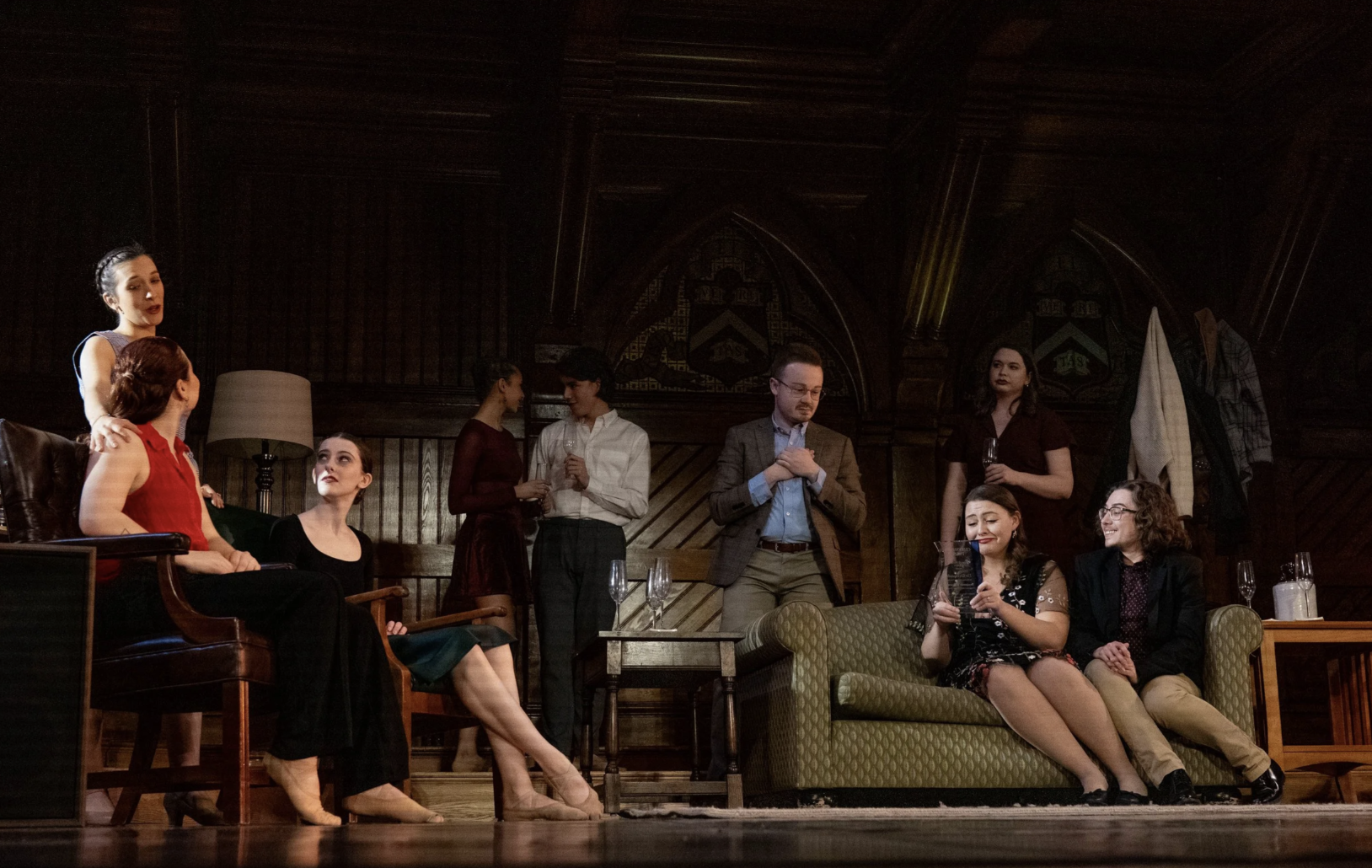A theatrical stage scene with multiple actors engaged in a play, set in a wood-paneled room with various furniture and props, including a lamp, a sofa, and chairs.