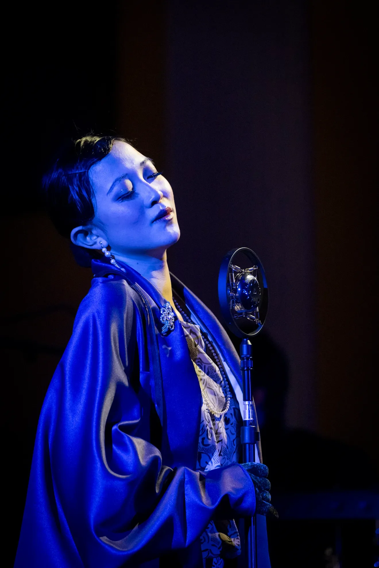 A woman with short dark hair dressed in vintage clothing, singing into a vintage microphone in a recording studio with dark background, illuminated by a blue light.