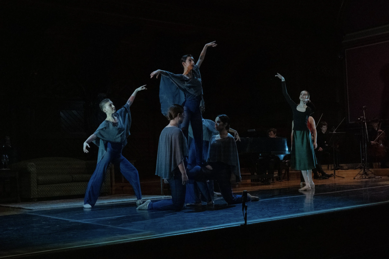 Ballet dancers perform on stage with one dancer balanced on the shoulders of two others, while an orchestra plays in the background.