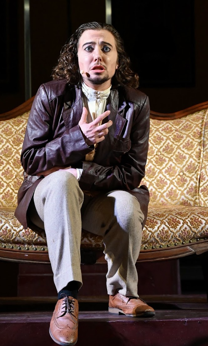 Person with long curly hair sitting on an ornate vintage sofa, wearing a leather jacket, beige pants, and brown dress shoes, with a surprised or confused facial expression.