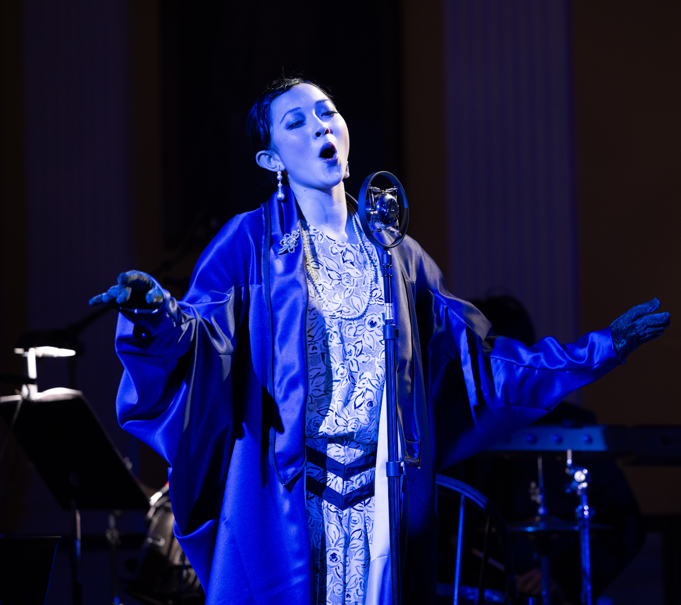 A woman singing into a microphone on stage, dressed in vintage clothing with gloves, illuminated in blue lighting.