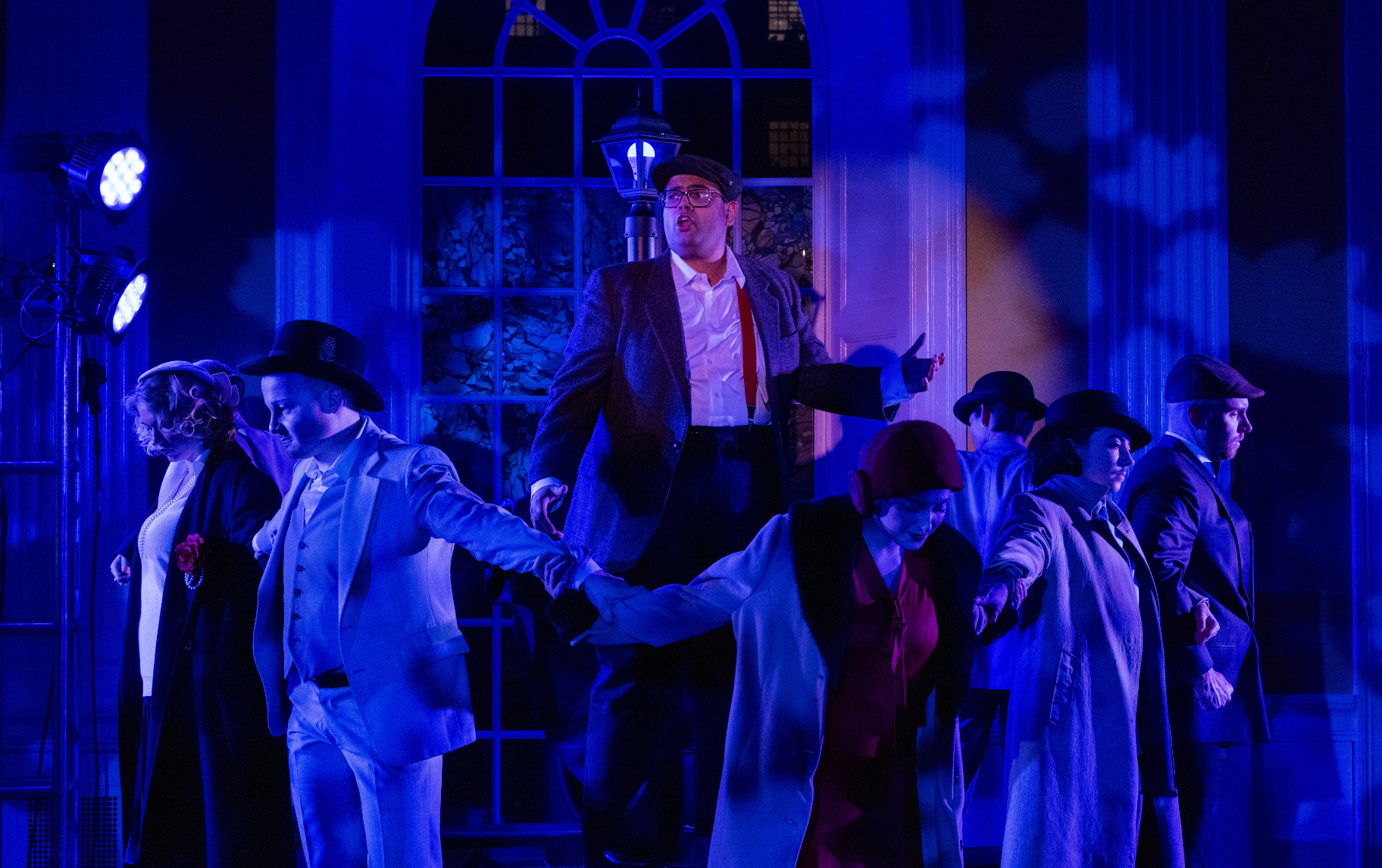 A stage performance featuring a man in a gray jacket and glasses, standing on a platform holding a phone. Six other performers, wearing vintage clothing with hats, are holding hands in a line at the front of the stage. The scene is illuminated with b