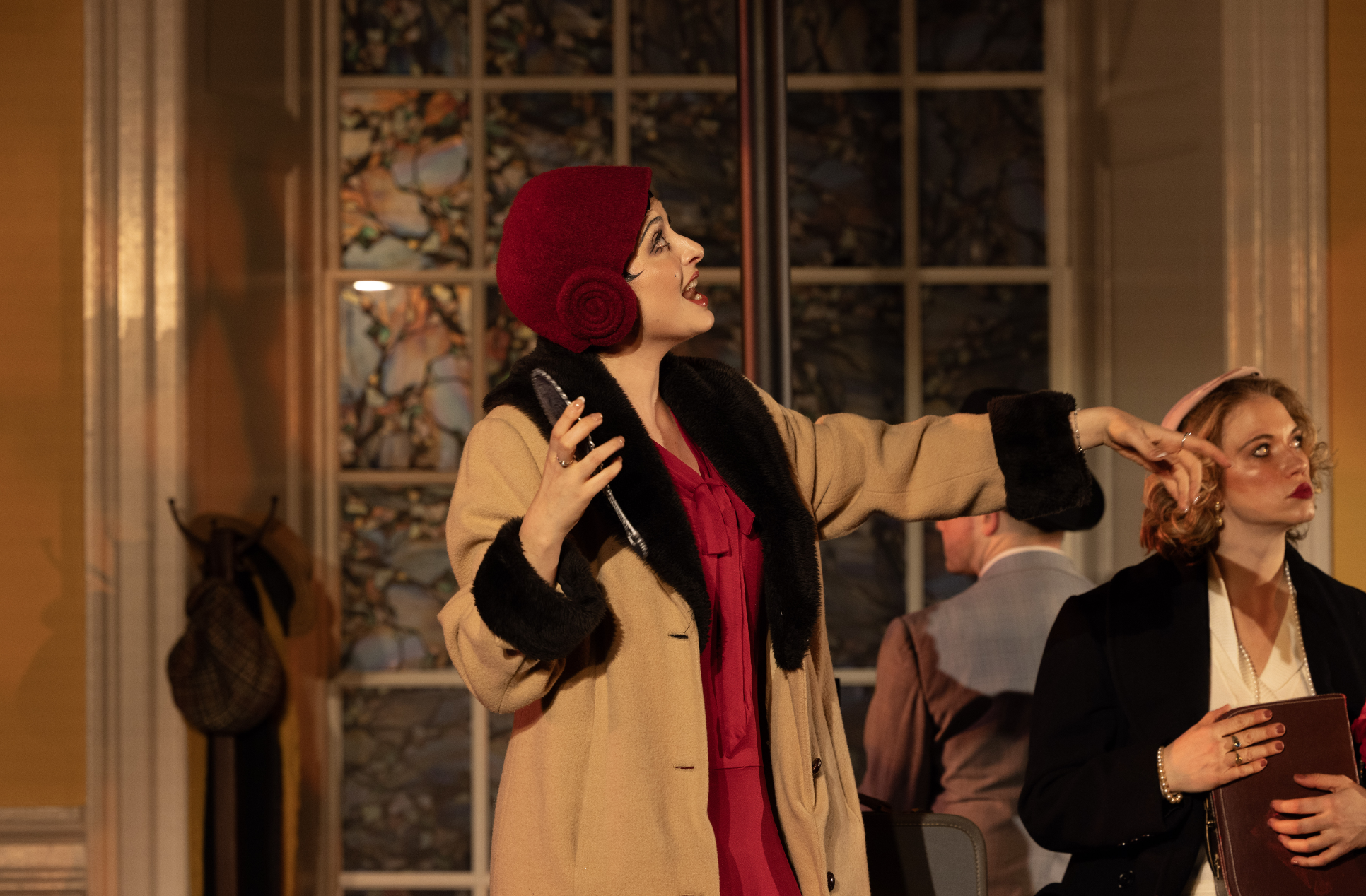 A woman dressed in vintage clothing, including a red hat and beige coat with black fur trim, gesturing and speaking at an indoor event with other people present.