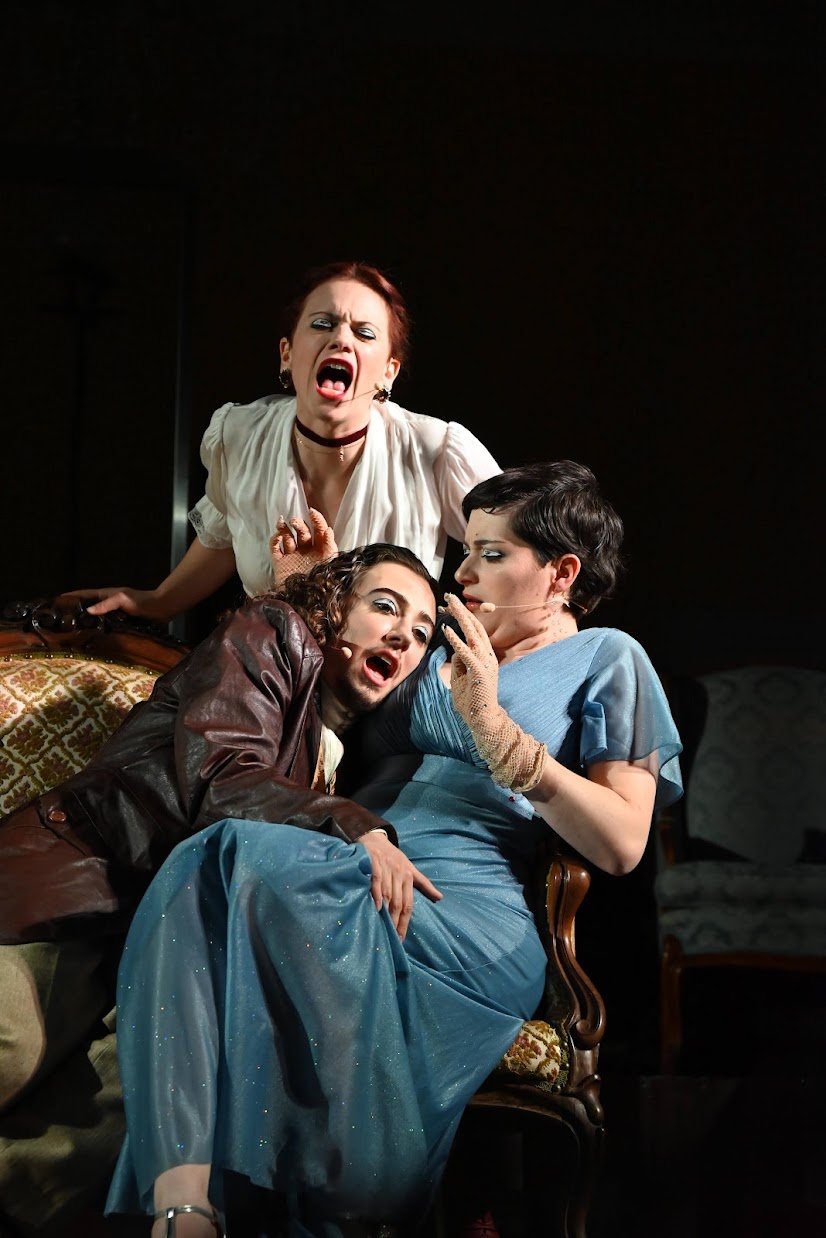 Three people acting dramatically on stage, with two women and one man, wearing theatrical costumes and makeup, engaging in an intense scene.