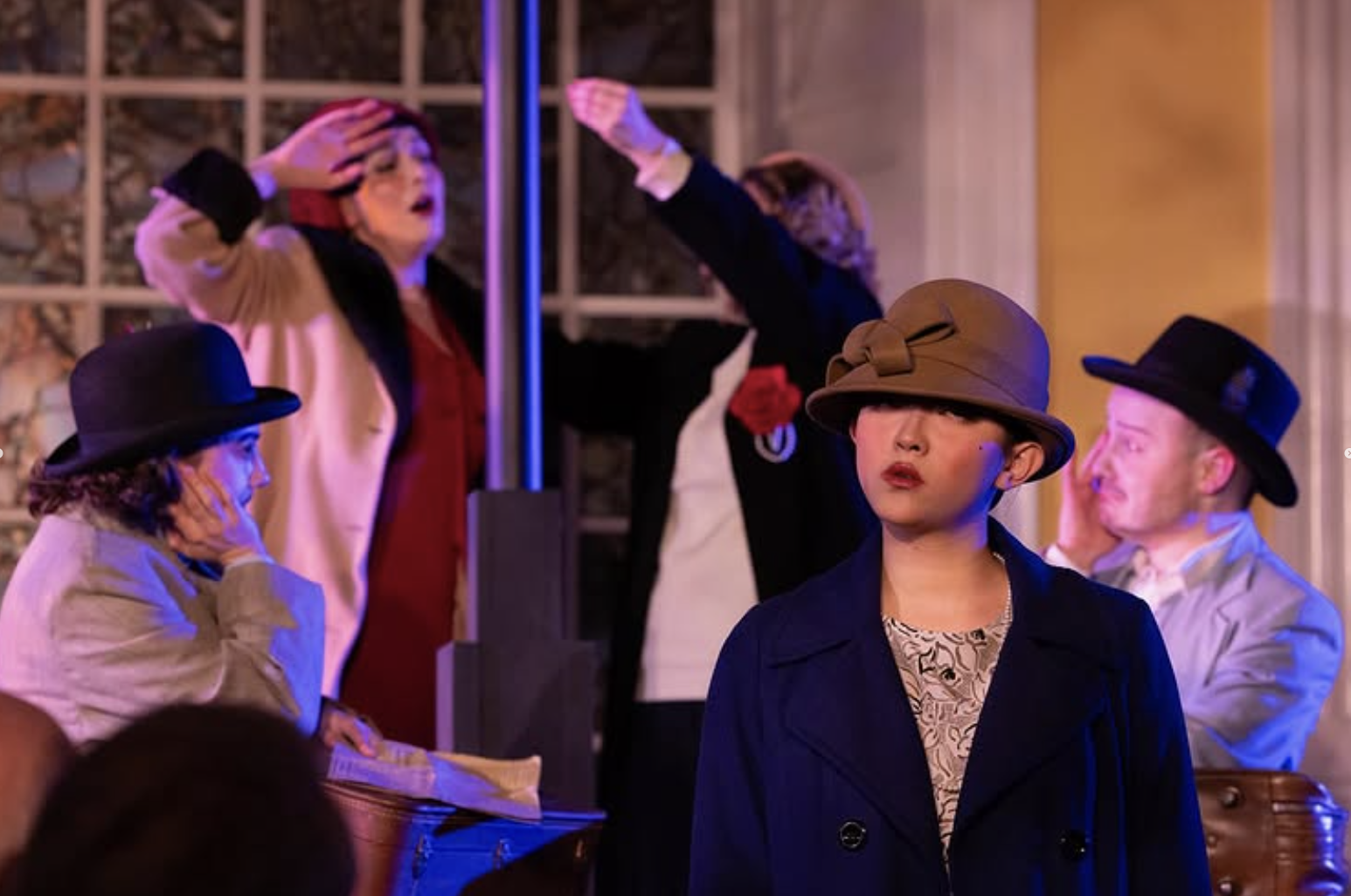 A group of people dressed in vintage attire, some wearing hats, at what appears to be a dance or performance venue with large windows in the background. One woman in the foreground has a serious expression and is wearing a brown wide-brimmed hat and 
