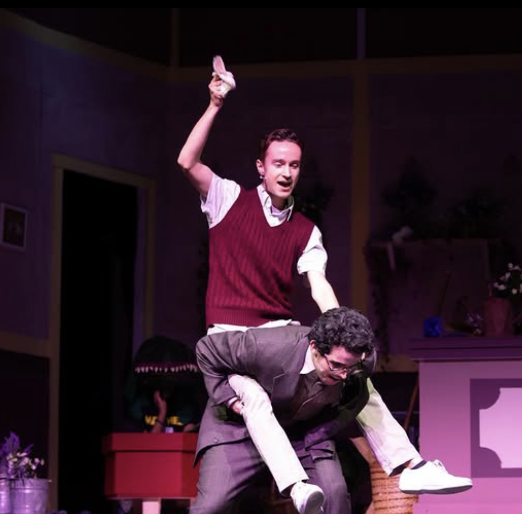 Two men performing a comedy scene on stage, one standing with one arm raised and the other crouching, wearing glasses and a suit, with a theatrical setting in the background.