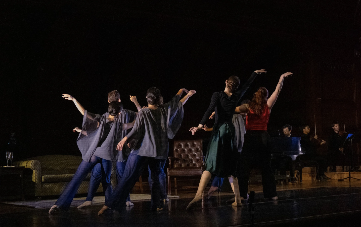 A group of ballet dancers perform on stage with dark background, some with arms raised gracefully, accompanied by musicians in the background.