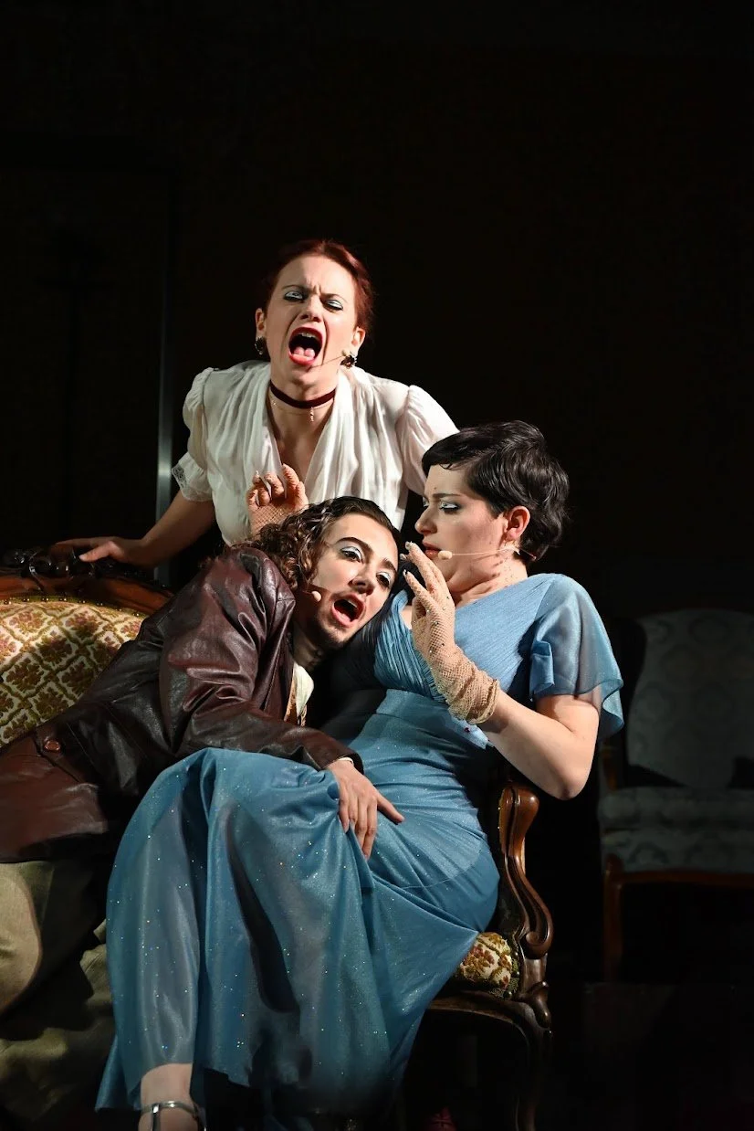 Three actors on stage. One woman standing behind, looks angry or shouting; a man sitting, appearing alarmed or surprised, with his head leaning on the woman’s shoulder; another woman seated, holding the man with a worried expression.